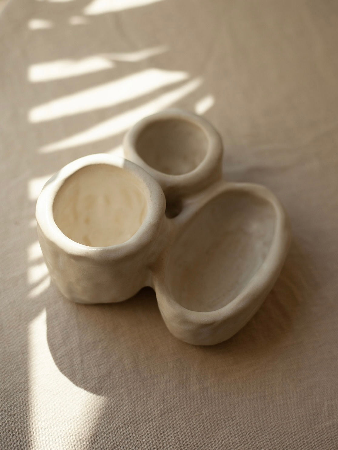 The Ceramic Plate 'Organic Fusion N°3' from INI Ceramique, a minimalist and organically shaped ceramic dish with three connected compartments, sits on a beige fabric surface. This stoneware piece is bathed in soft natural light, casting gentle shadows that highlight its smooth, uneven texture.