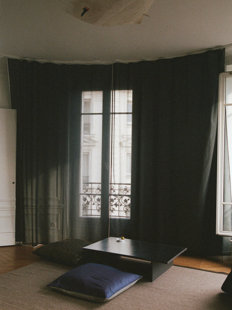 In a minimalist room with large dark curtains partially open to reveal a balcony and window, a low Mara Sister Coffee Table – Silver by TEGET sits on a beige rug surrounded by dark cushions. Light streams in, crafting a cozy and serene atmosphere.