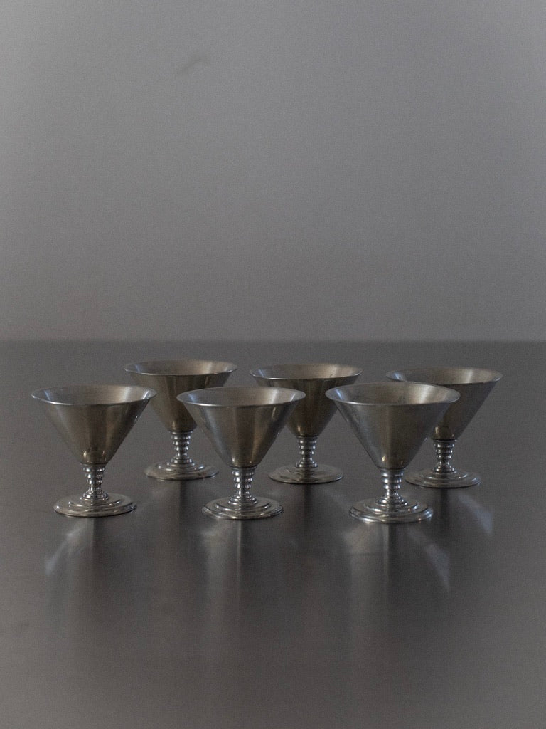 A set of the "Six Ice Cream Cups" by Out For Lunch is evenly spaced and arranged in two rows on a reflective surface against a plain grey background. These small tin goblets, reminiscent of vintage ice cream cups, feature sleek, conical shapes on short stems, evoking a touch of Scandinavian elegance with their minimalist design.