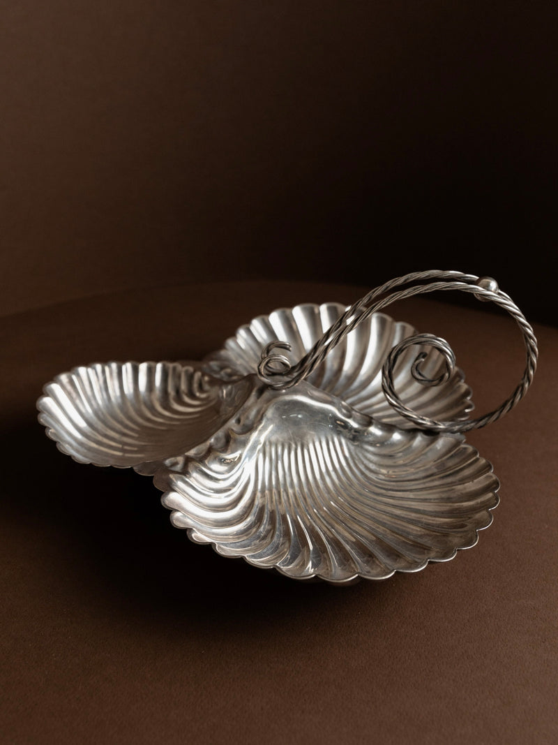 The "Silverplated Centrepiece Shell Shape" by Bottega Jacobs features three shell-shaped compartments and an ornate, curved handle, making it a stunning vintage centerpiece against a dark brown background.