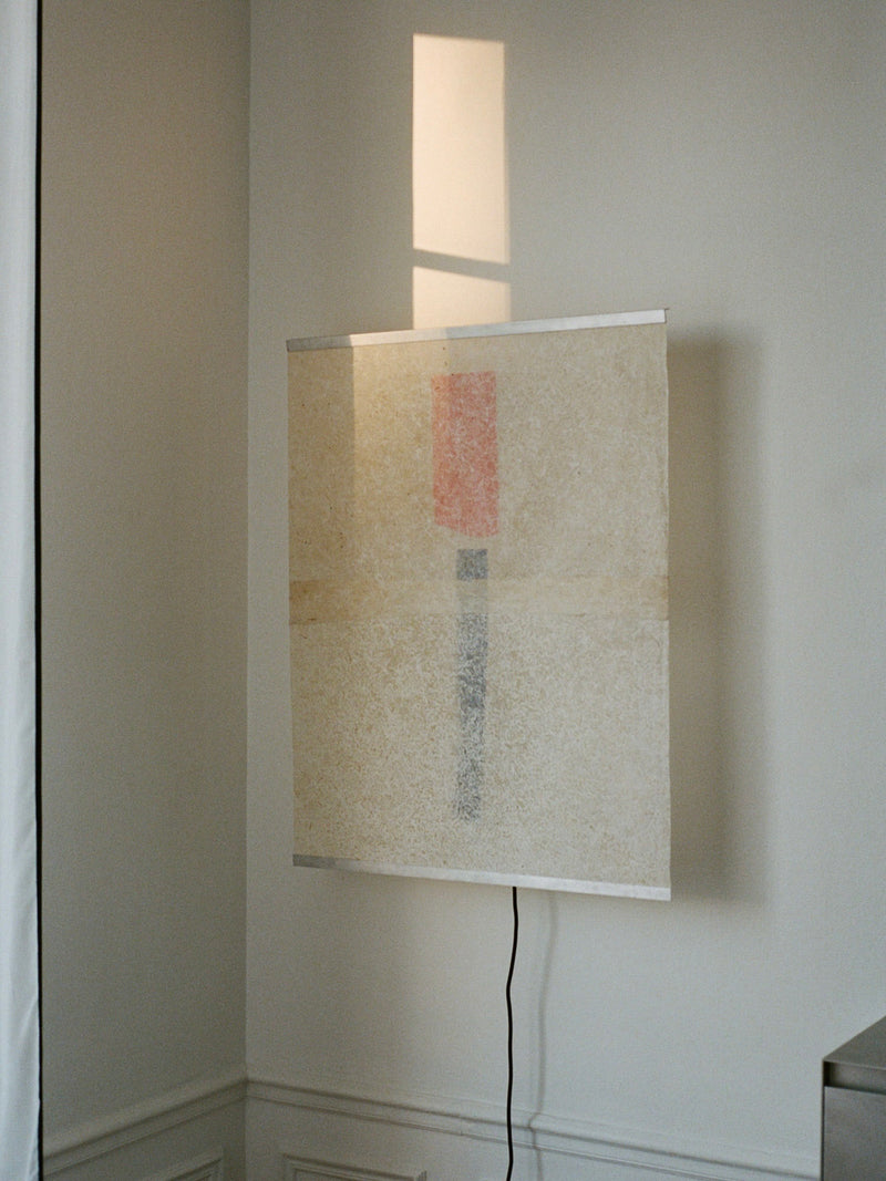 The Panel Lamp – Esei by Ana Kraš for TEGET hangs on a white wall, displaying a minimalist design with red and blue segments in a vertical line. Illuminated softly by natural light, it casts gentle shadows. A black cord, echoing a wall lamp's utility, complements the artwork's elegance.