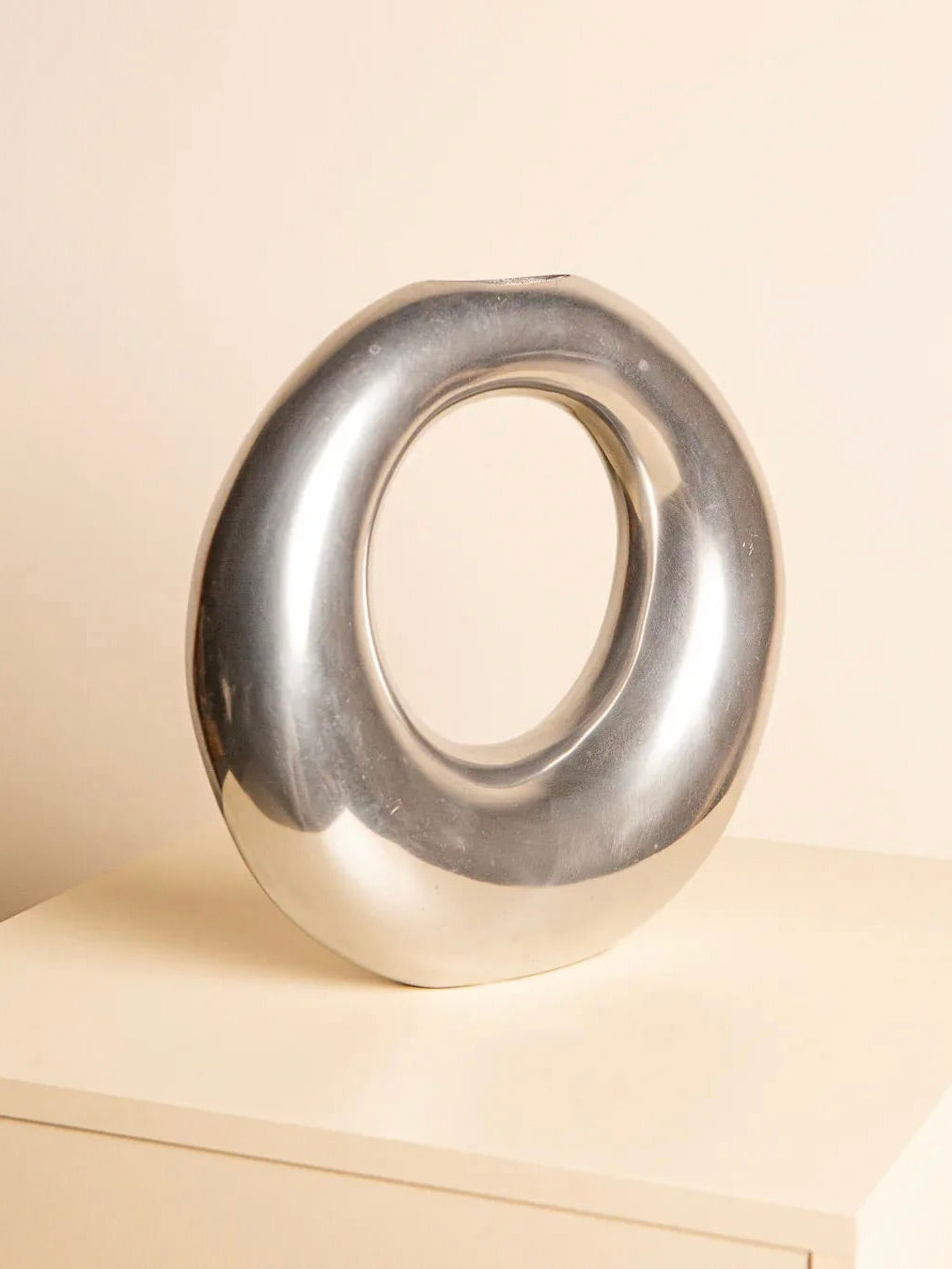 The Treaptyque Aluminum "Sac" Vase, a shiny silver metallic sculpture with a modern, circular hollow design, rests on a light-colored surface against a neutral background. Its smooth and reflective surface beautifully catches the ambient light.