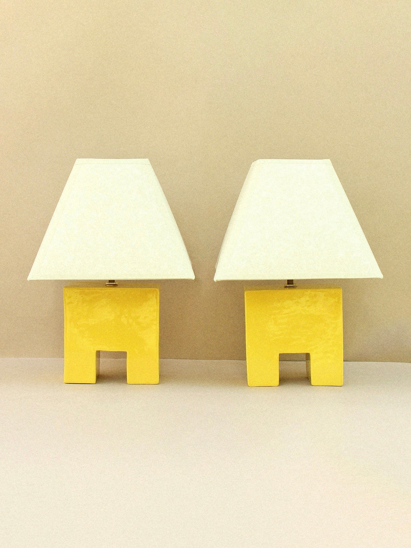 Two Cristo Lamp (Sol) by Casa Veronica, featuring yellow ceramic bodies and custom white trapezoidal shades, are placed side by side against a neutral beige background.