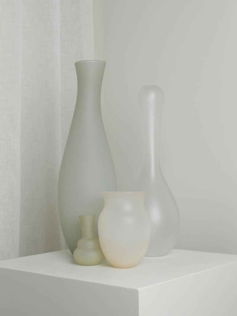 Four frosted glass vases of varying shapes and sizes, including the Medium Vase in Sahara by Verre d'Onge, stand on a white pedestal against a neutral background with soft, sheer curtains on the left. Mouth-blown and minimalistic in design, the vases showcase hues of white, beige, and light grey reminiscent of Sahara sands.
