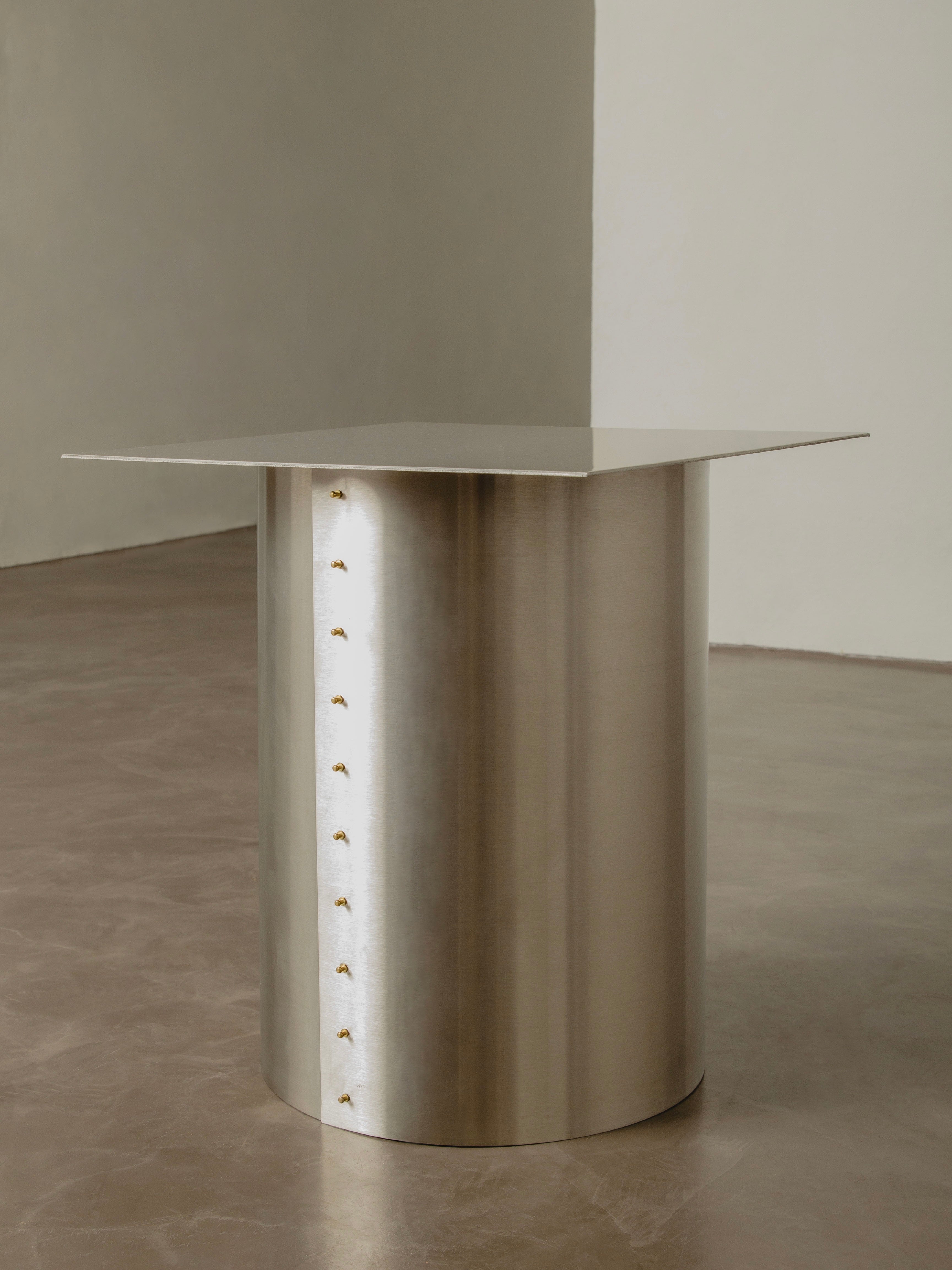 Introducing the "Plataforma" Side Table by Pedro Avila, a sleek and modern piece that features a rectangular top and a cylindrical base crafted from polished metal, reminiscent of astronomical instruments. The base is elegantly adorned with a vertical row of small decorative studs, making it an epitome of minimalist elegance from the technological era. It stands beautifully on a smooth brown floor against a neutral wall.