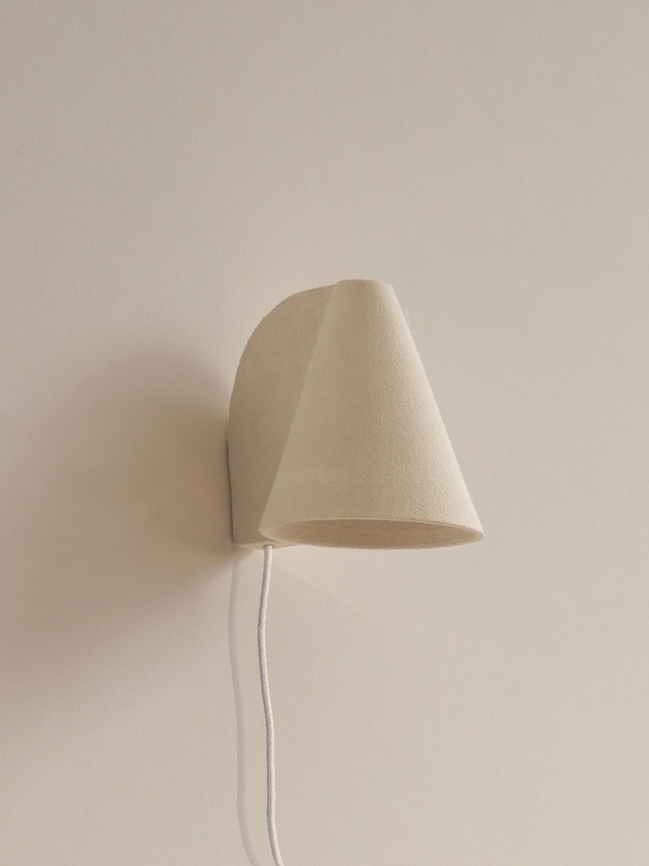 A minimalist wall sconce by Mariza Galani, the Esoteric Wall Lamp features a beige conical lampshade made from a softly textured material, with its white cord elegantly draping down against a light, neutral wall.