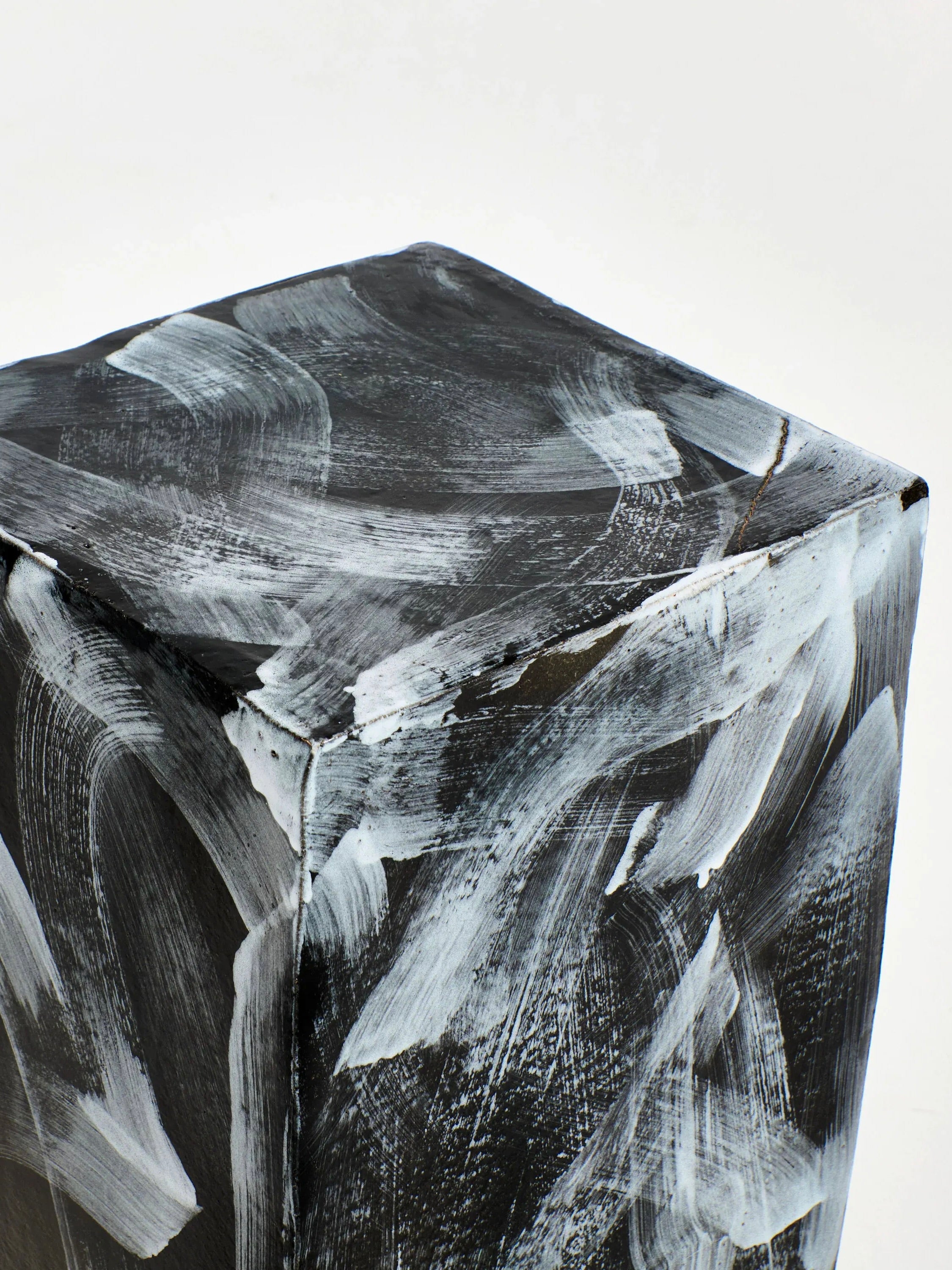 The Ceramic Side Table, Geometric Shape from Project 213A is a textured cube sculpture with a predominantly black surface featuring bold white brush strokes in an abstract pattern. Handmade in Portugal, this piece possesses visible imperfections and a rough, artistic finish, presented against a plain white background.