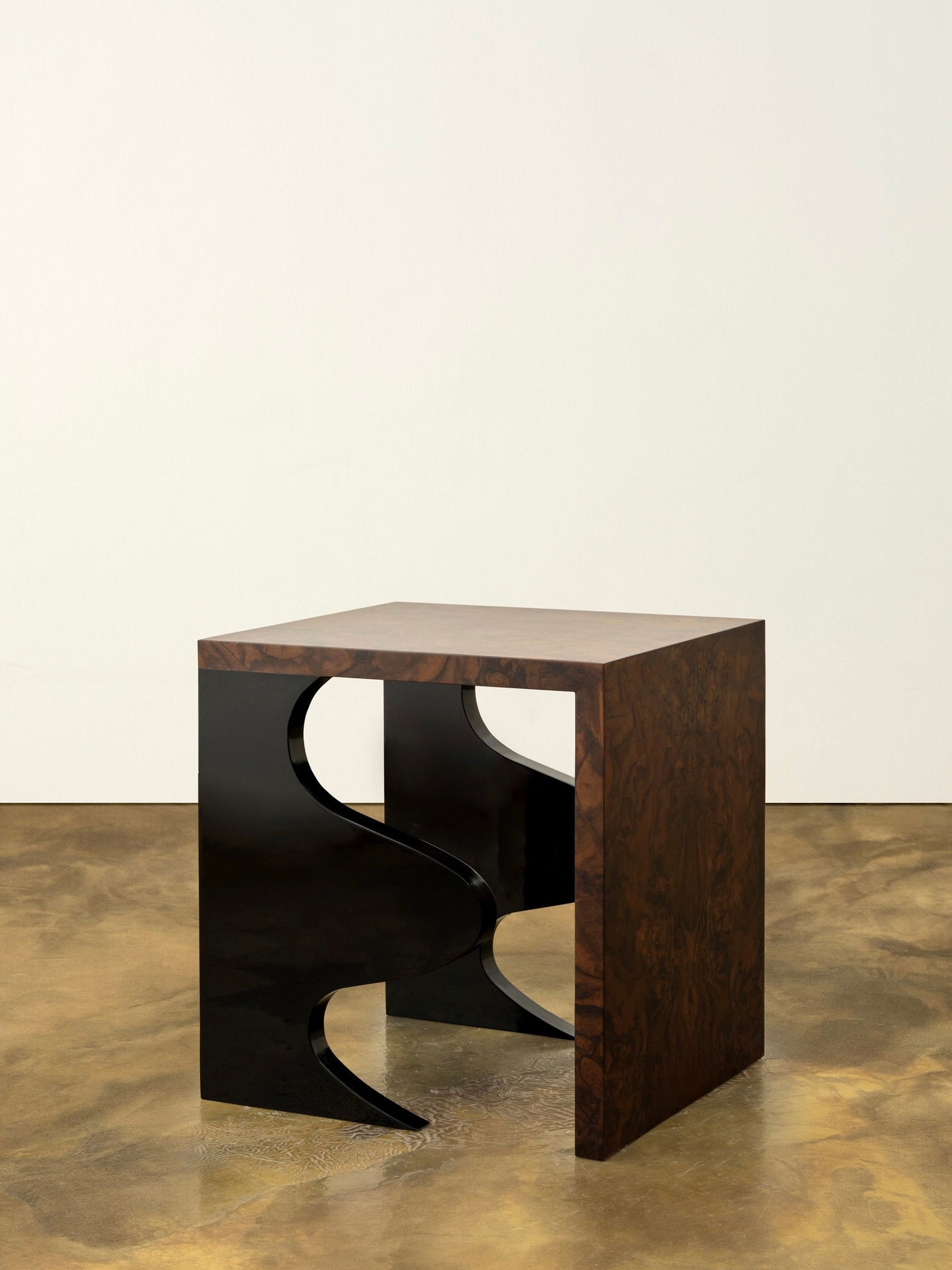 The Harlequin Side Table by Animate Objects features a walnut burl veneer square top with abstract black metal legs. Its glossy lacquered wood finish stands out magnificently against the polished brown and gold floor, while the plain white wall creates a striking backdrop for this luxurious piece of furniture.