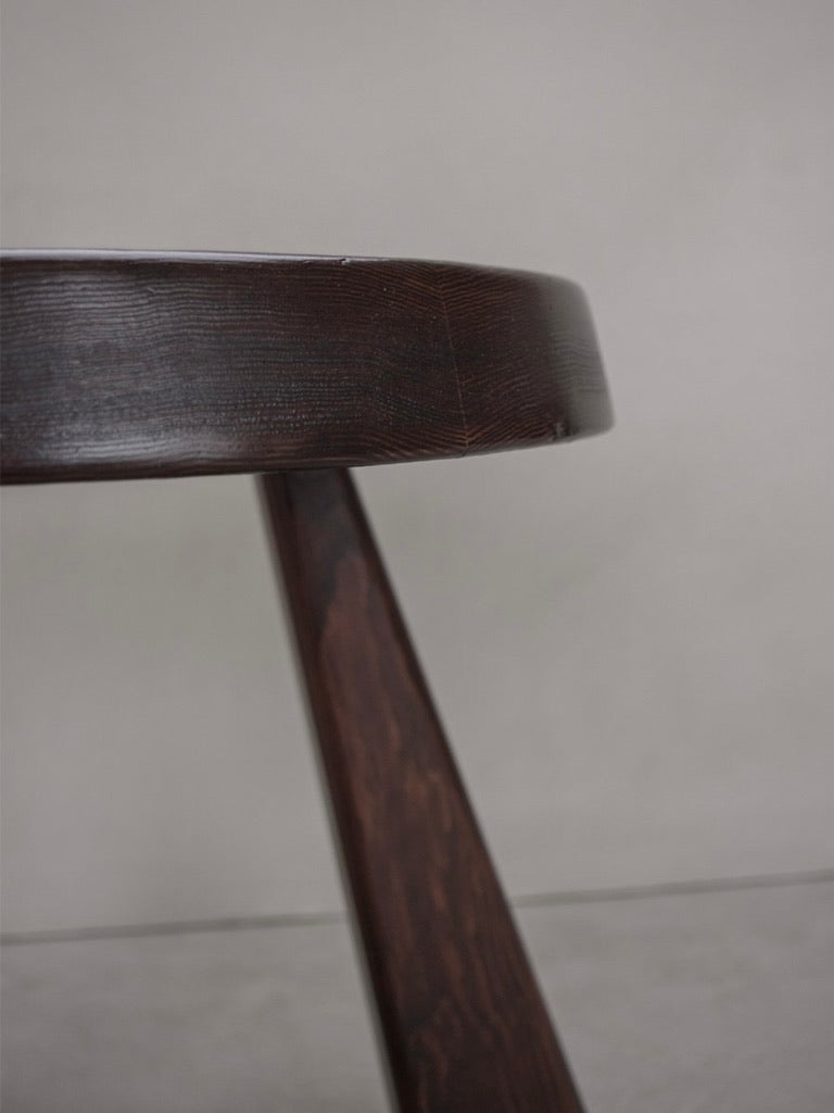 Close-up of a Bicci de' Medici Charlotte Perriand Sandoz Stool, featuring a round edge and elegantly crafted single leg in rich mahogany wood, beautifully contrasting against a plain, light gray wall.