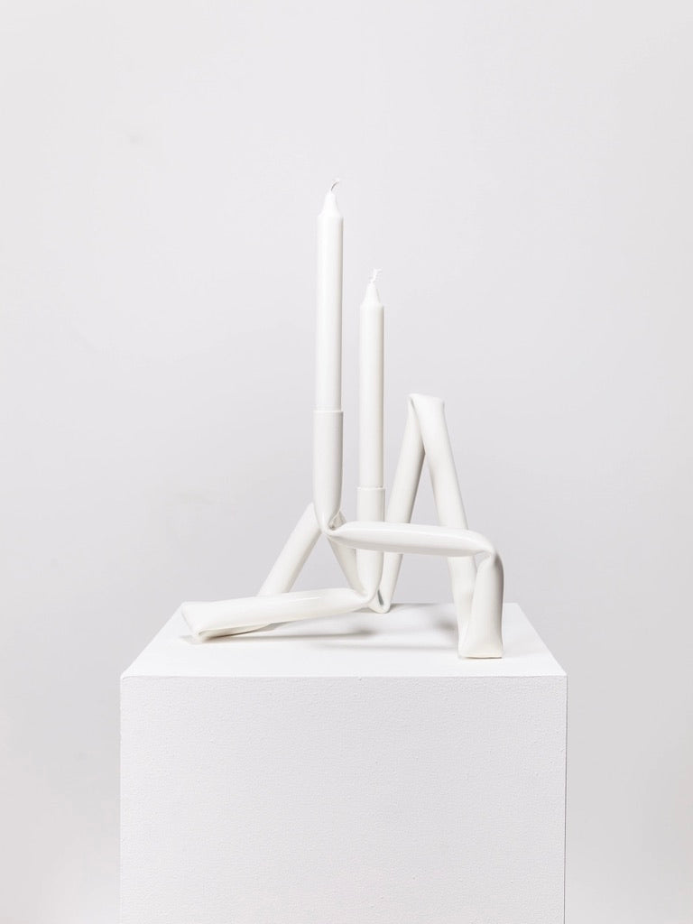 The Bucatini candle holder (White) from AOT STUDIO's Primi collection is a minimalist, hand-crafted piece featuring a unique, abstract design with intertwined, angular shapes made from aluminum tubing. It elegantly supports two unlit white candles of varying heights and is displayed on a white pedestal against a plain white background.