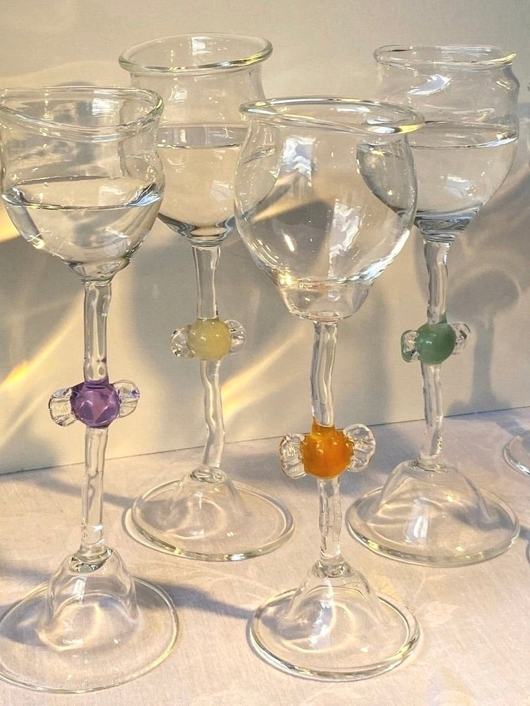 The Mouth Blown 'Candy' Wine Glasses by Vivian Alarcón feature four clear glasses, each with a colored orb—purple, yellow, orange, or green—on the stem. These vintage pieces are handcrafted and stand elegantly in natural light.
