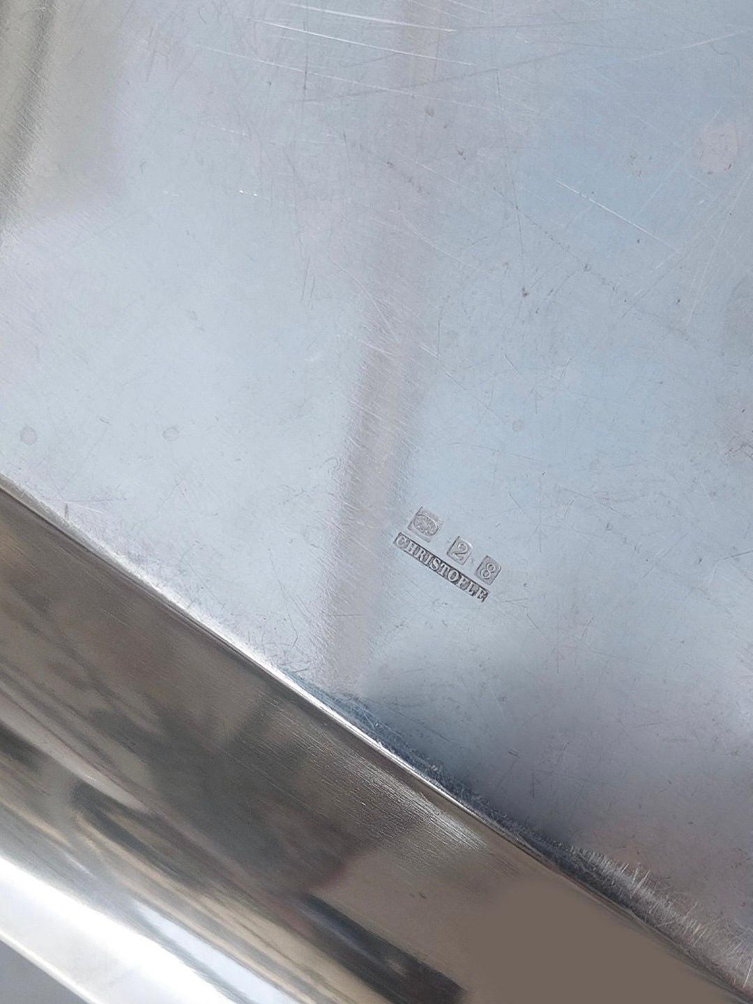 A close-up view of the polished surface of an Octogonal Christofle Dish by Les Objoies, featuring a hallmark with three small symbols and the text "STAINLESS STEEL." The surface exhibits slight scratches and reflections, suggesting it is a frequently used piece of elegant tableware.