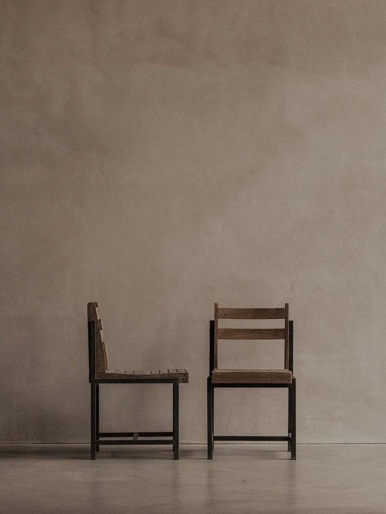 Two Bicci de' Medici Pierre Jeanneret Authentic PJ-SI-65-A chairs with metal frames and teak slats sit in a minimalist room with a beige wall and light floor. One chair is forward-facing, the other sideways, creating an atmosphere of simplicity and calmness.