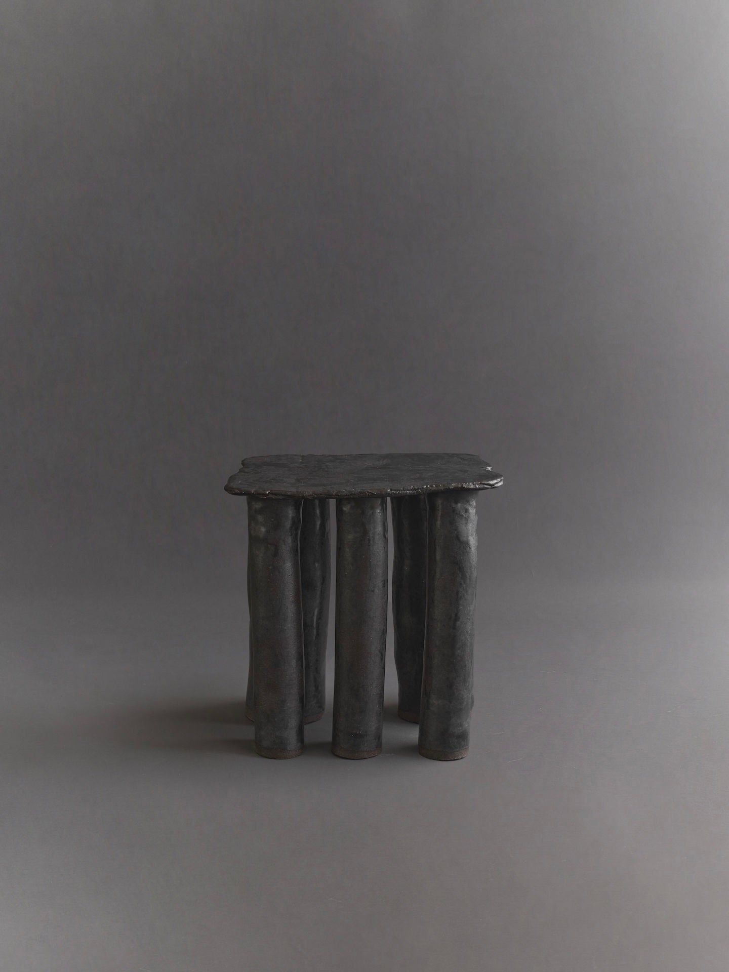 The Cleo Ceramic Side Table by Ombia Studio is a small, minimalist black table with a rectangular top and uneven cylindrical legs. It features a rustic, handcrafted look enhanced by a textured black glaze, set against a plain gray background.