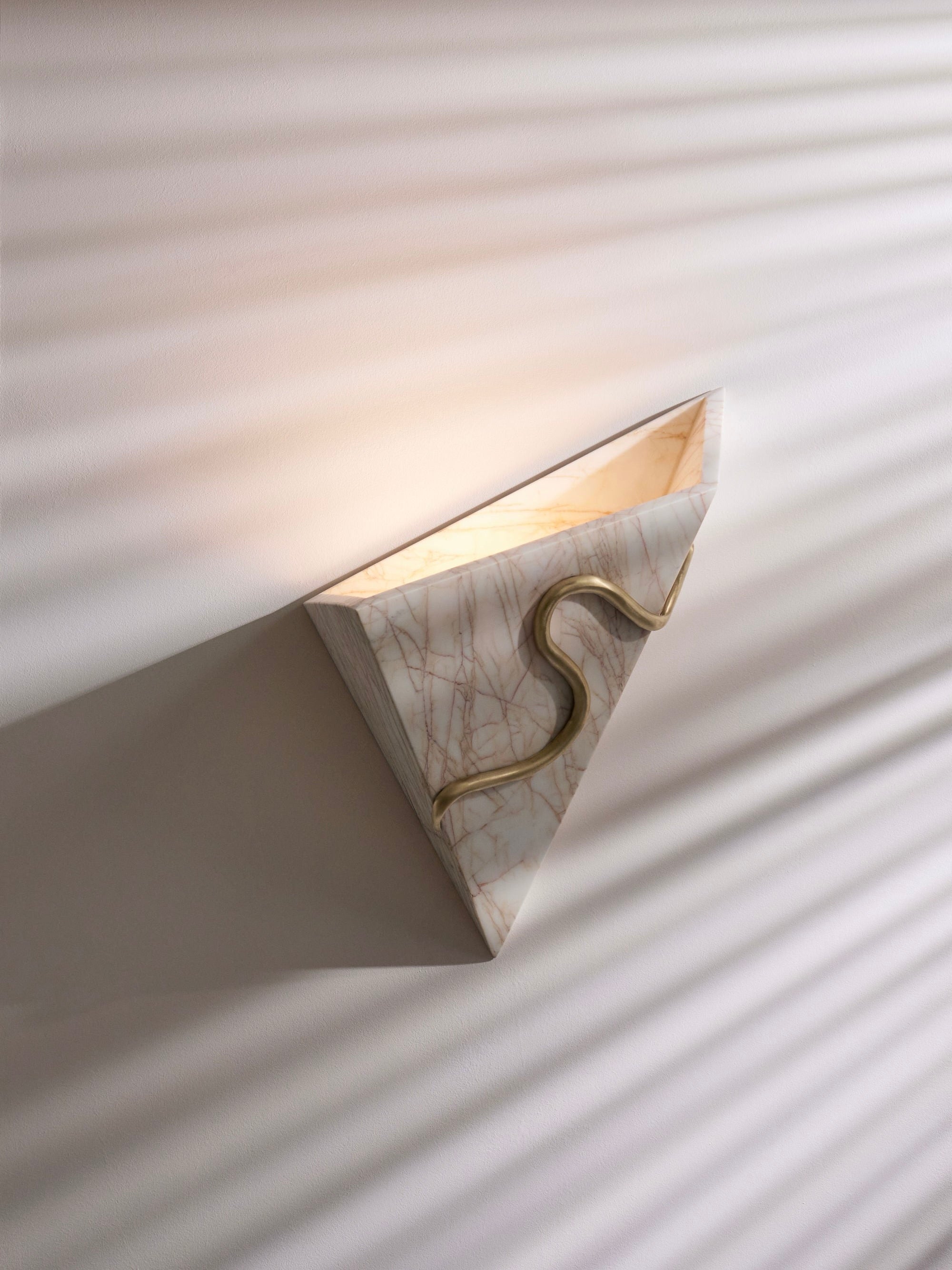 The Therapeutic Wall Light 2 by Mariza Galani is a triangular wall sconce crafted from marble, featuring a golden curving design in the middle that emits soft, therapeutic light. The shadows cast from this handcrafted fixture create striped patterns on the wall, enhancing its minimalist and modern aesthetic.