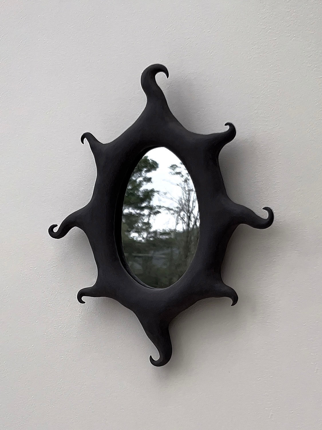 A Black Sun Mirror by Solenne Belloir, boasting an oval shape with a matte black finish and an intricately wavy frame reminiscent of stylized flames or sun rays, is mounted on a pale wall. The mirror reflects an outdoor scene featuring trees and a cloudy sky, infusing the room with a magical presence.