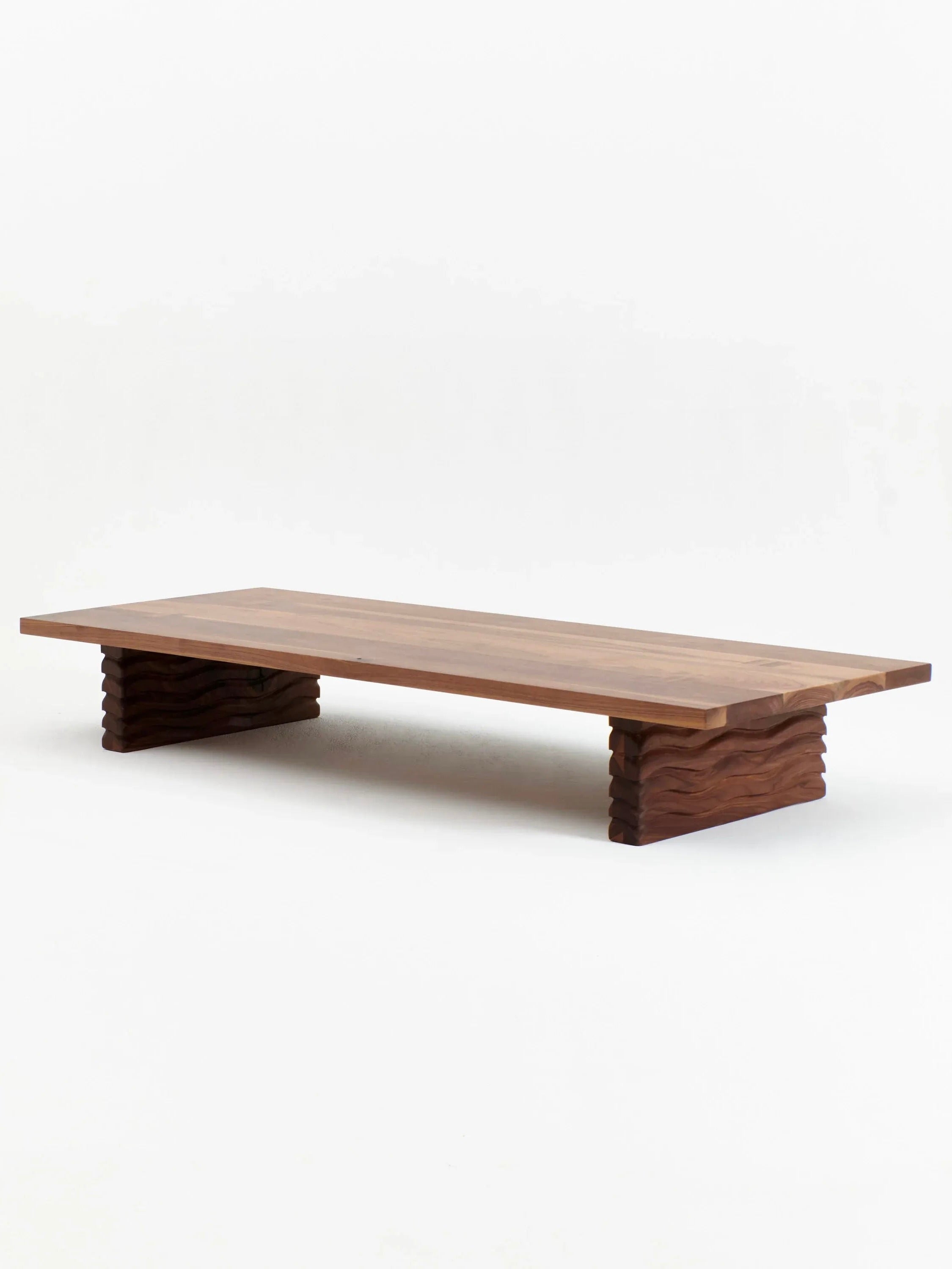 The Nazaré Daybed by Project 213A is a low wooden furniture piece featuring a rectangular top and two wide, blocky legs with a wavy, textured pattern. Handmade in Portugal from solid walnut, this minimalist daybed showcases a natural wood finish that beautifully highlights the grain and color variations of the wood.