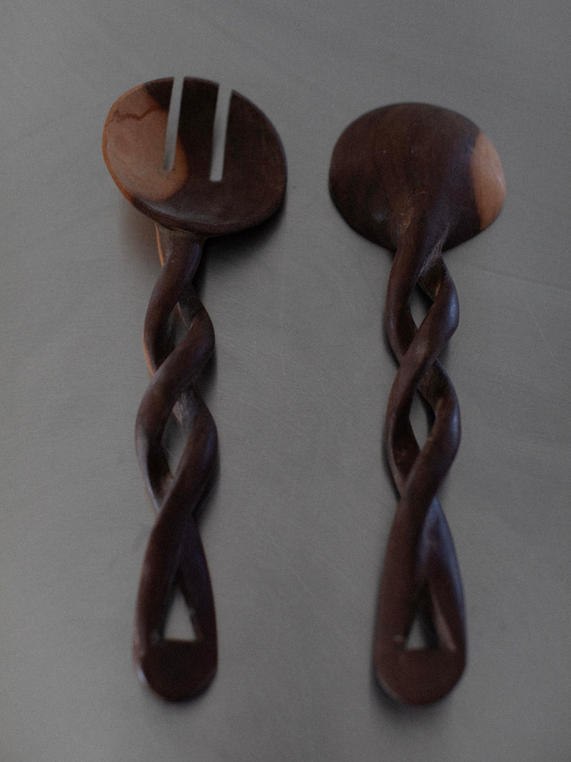 A set of two Twisted Serving Spoons from Out For Lunch lies on a flat surface. One wooden salad server has a slotted end while the other has a solid end. Both utensils, made through an intricate hand-carving process, feature twisted handles and are polished to reveal their natural grain.