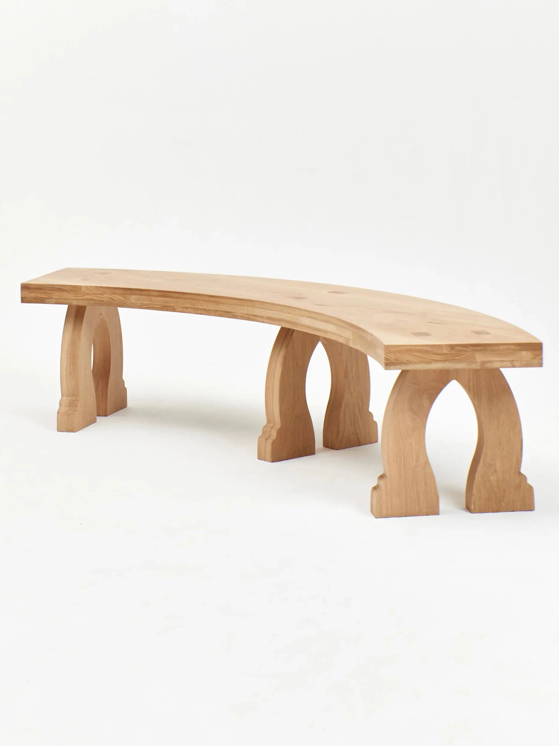 The "Sintra" Curved Bench in Oak by Project 213A features a slightly curved seat and four chunky, arch-shaped legs. Crafted from premium oak wood by expert artisans, this bench boasts a natural wood finish and minimalist design, standing elegantly against a plain white background.