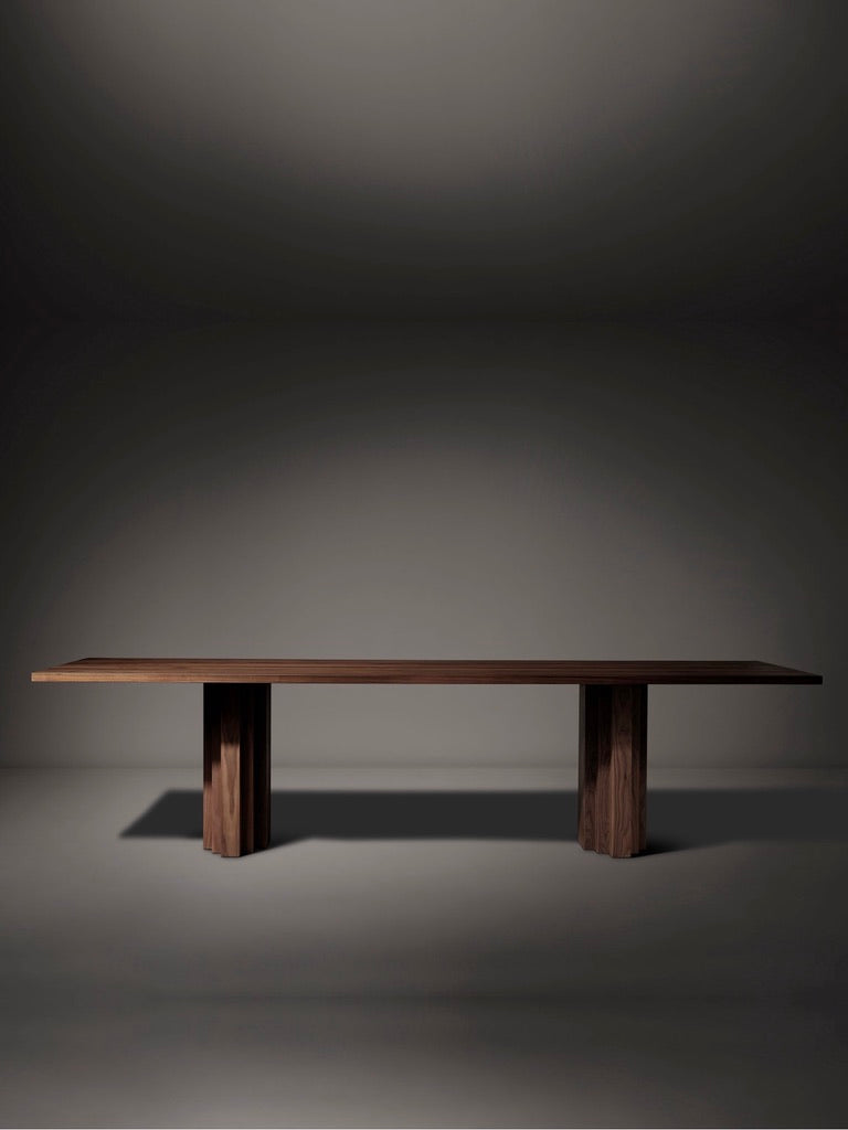 The Mokko Brut Slim Table, made from dark American walnut, features a minimalist design with two sturdy block legs. Displayed against a softly lit plain background, it radiates elegance and simplicity.