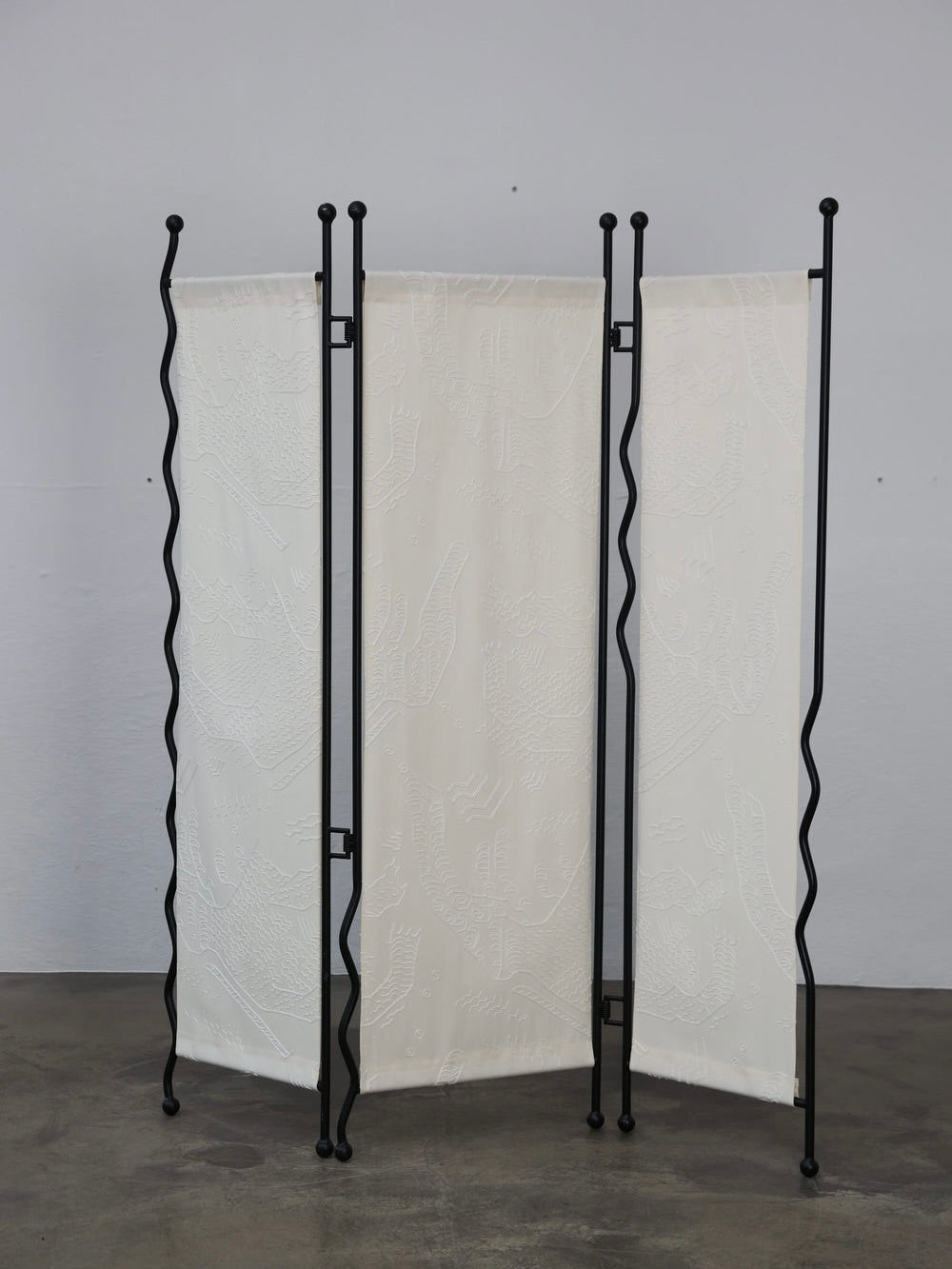 The Good Living&Co.'s "Alto" Folding-Screen features black metal frames and white fabric panels with a unique embossed pattern, elegantly sitting on a smooth gray floor for both style and function.