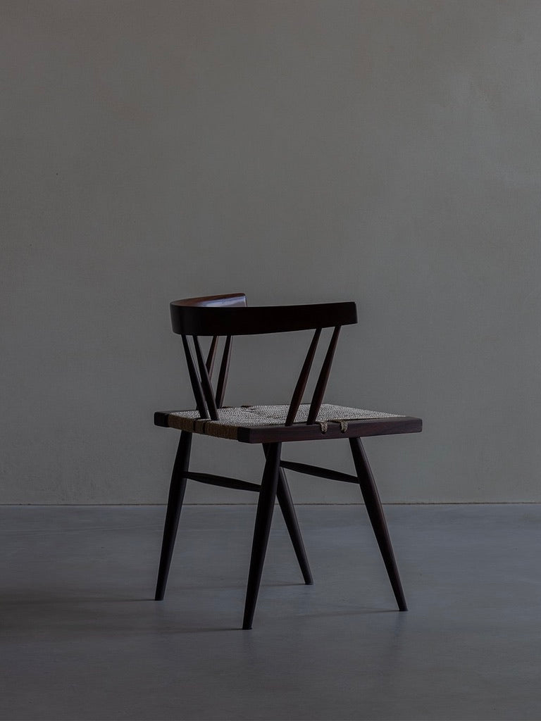 The Manner of Nakashima Grass Seated Chair by Bicci de' Medici, with its minimalist rosewood design, angled legs, and low backrest, evokes mid-century allure. It's positioned in a dimly lit room against a plain gray wall, reminiscent of George Nakashima's modern aesthetic.