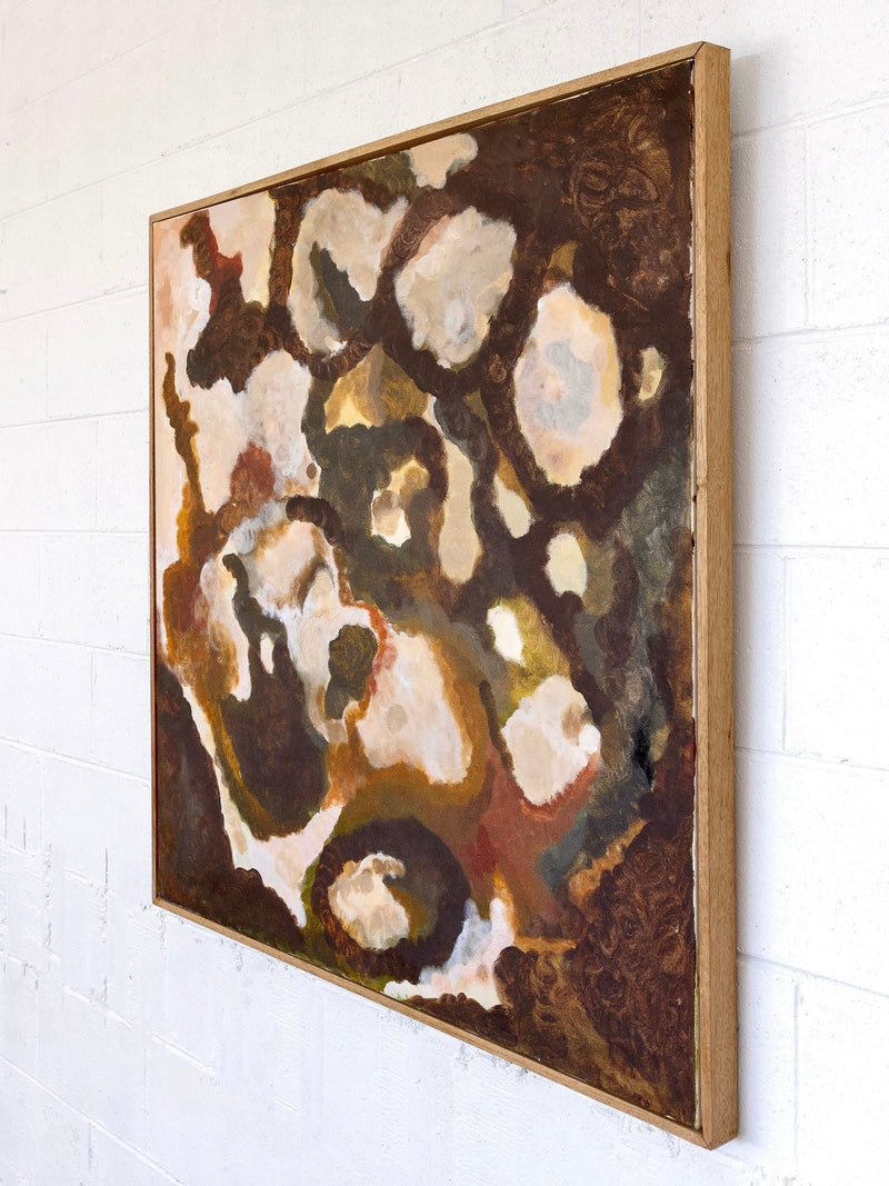 A side view of "That Which Remains," a large abstract painting by Natasha France, mounted on a white brick wall. The artwork, rendered in mixed media on canvas, features swirling, cloud-like shapes in earthy tones of brown, beige, and white. The painting is framed in elegant Tasmanian Oak.