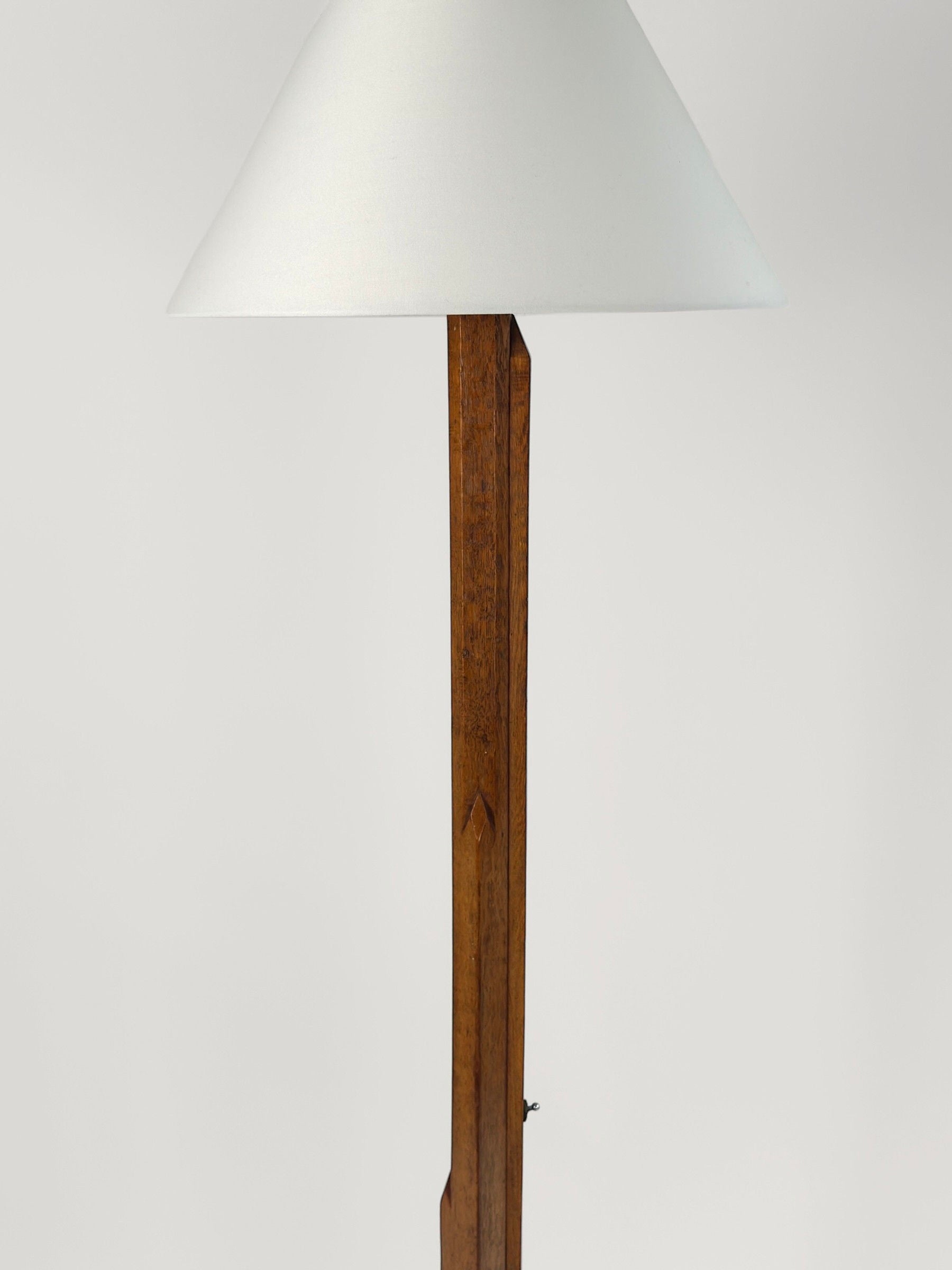 Introducing the Art-Deco Floor Lamp by Figuresdesign: a wooden floor lamp with a sleek, angular design and a white, cone-shaped lampshade. The lamp features a tall, rectangular carved wood base with clean lines and a natural finish, standing against a plain white background. This elegant piece evokes the timeless sophistication of an Art-Deco floor lamp from circa 1930.