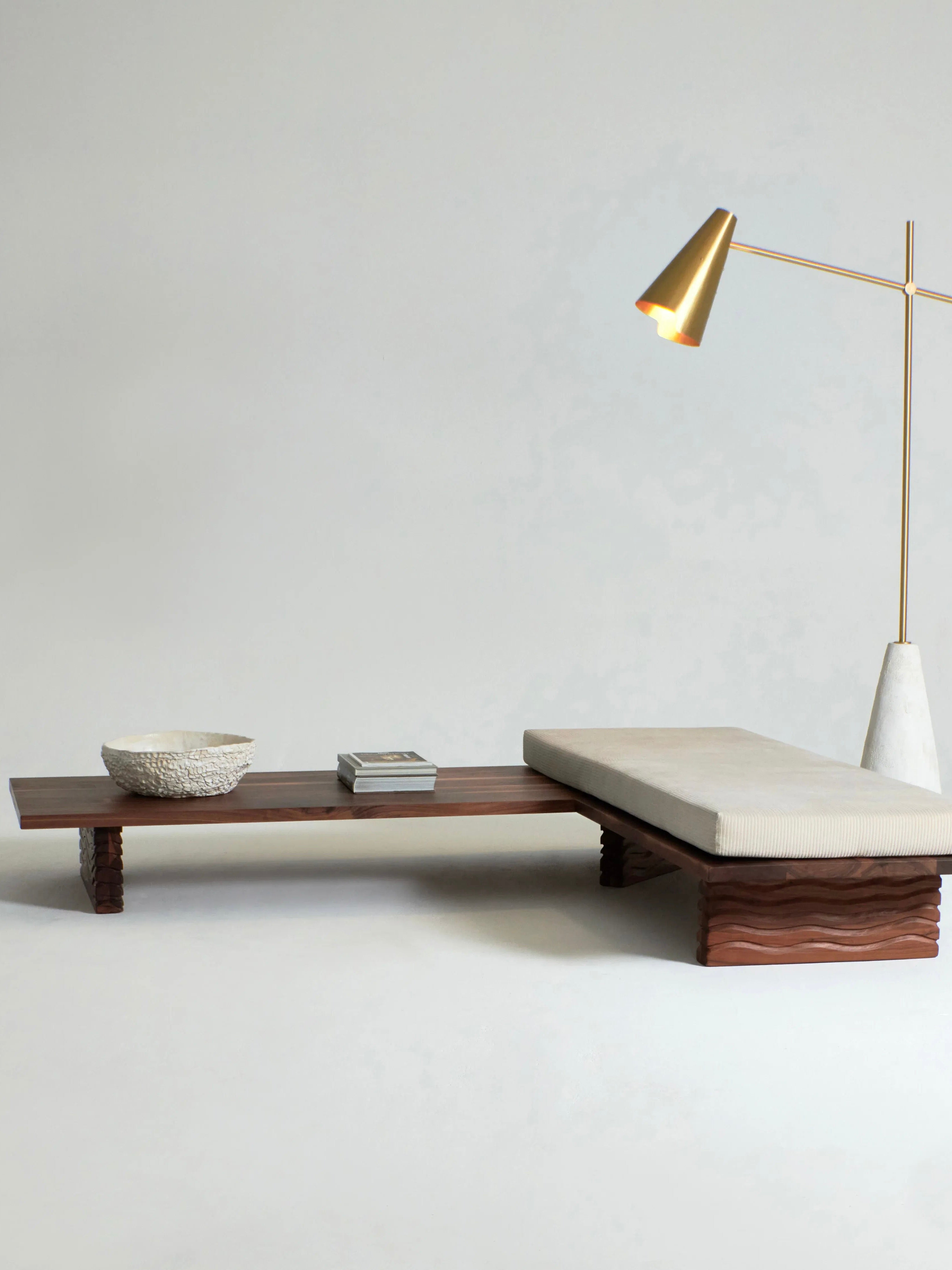 A minimalist room featuring the Nazaré Daybed L Shape by Project 213A, crafted from solid walnut with sculpted legs. The daybed has a removable cushion on one side and is paired with a textured bowl and a stack of books. Beside the daybed, a modern brass floor lamp with a conical shade casts light over the setup against a plain backdrop.