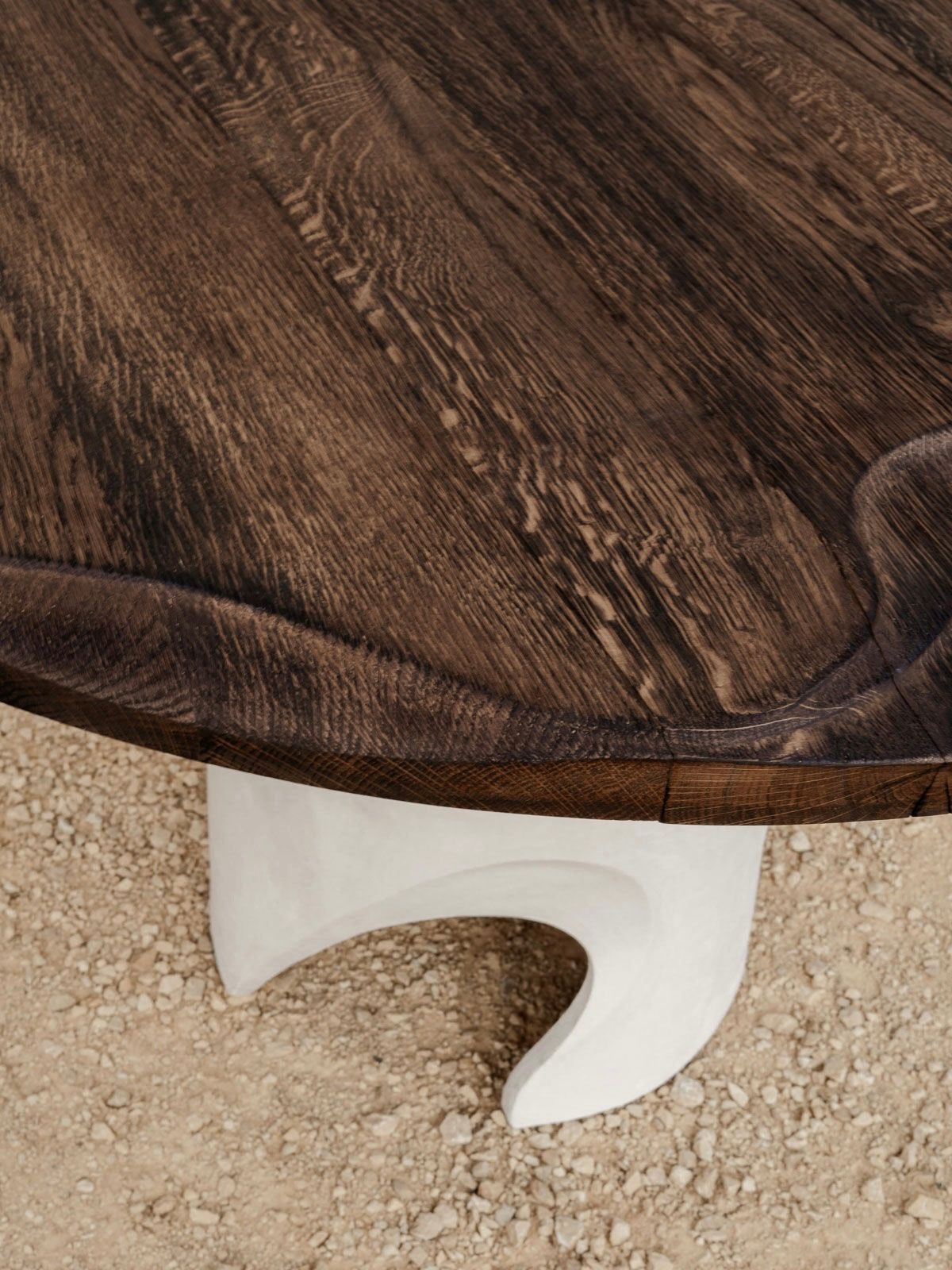 A close-up of Lea Bigot’s Lac - Carved Table in Sandstone and Oak reveals a round wooden top with dark, textured surfaces and sculpted grain, paired with a wavy white base showcasing modern design on gravel while highlighting natural details.