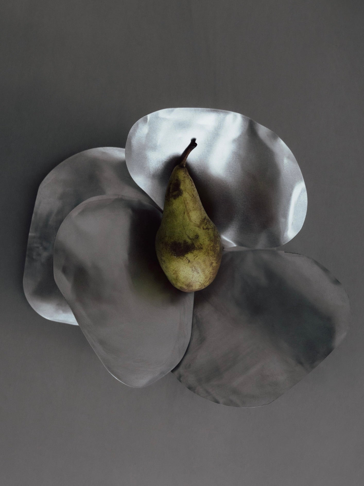 A pear rests on overlapping, irregularly shaped Petale Bowls with a reflective texture, set against a dark, neutral background. The stainless steel arrangement by The Good Living&Co. creates a contrast between the organic form of the pear and the smooth, shiny surfaces.