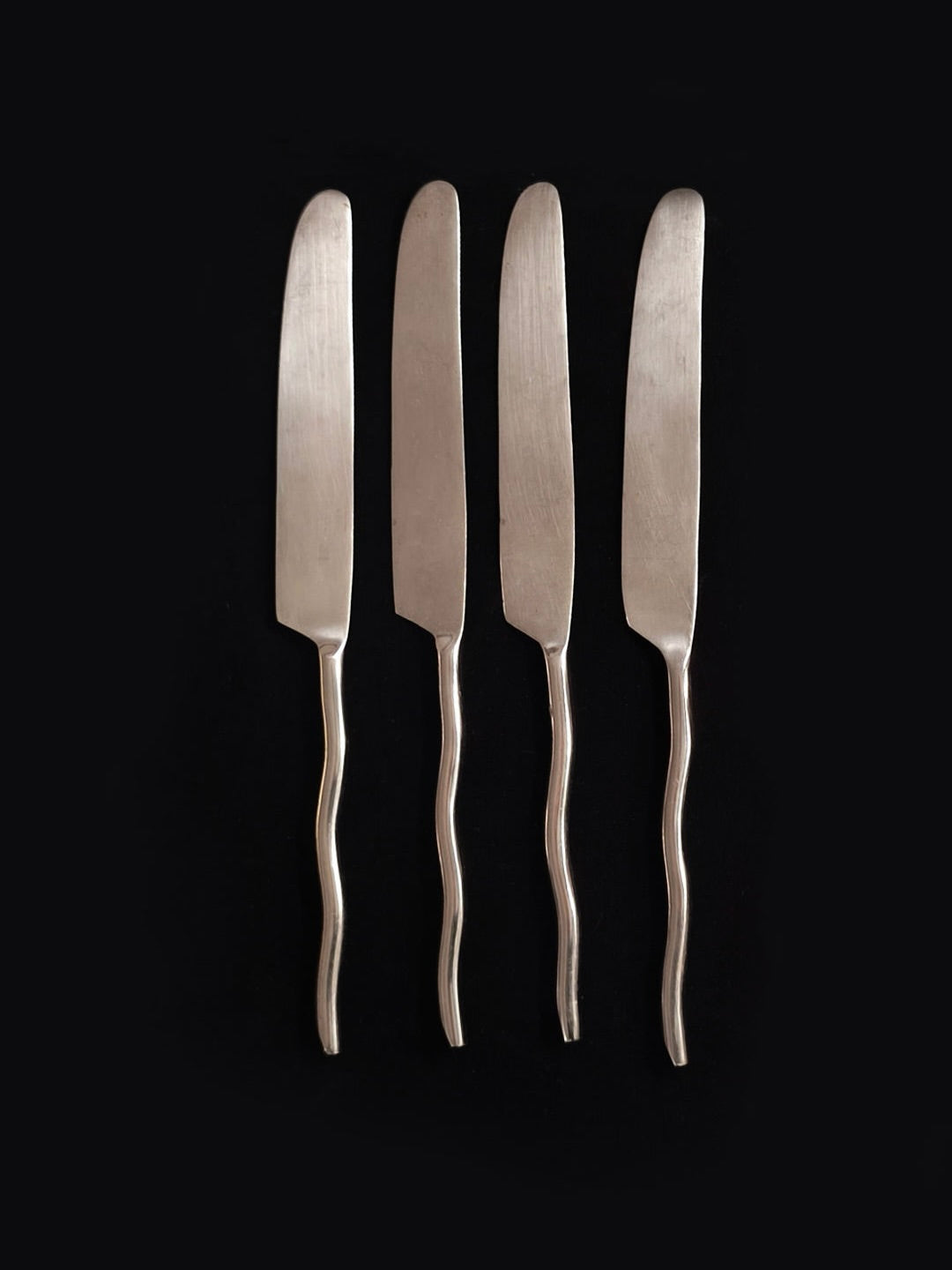 A set of four silver knives from Les Objoies, featuring elegant wave handles, arranged vertically against a black background.