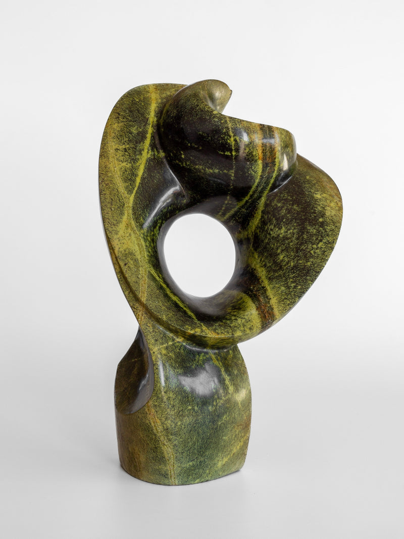 Featuring smooth, curved shapes with a circular opening in the center, the Abstract Stone Sculpture I by Spigel is crafted from leopard stone by a Zimbabwean artist. Its surface showcases a dynamic blend of green and black hues, all set against a plain white background.