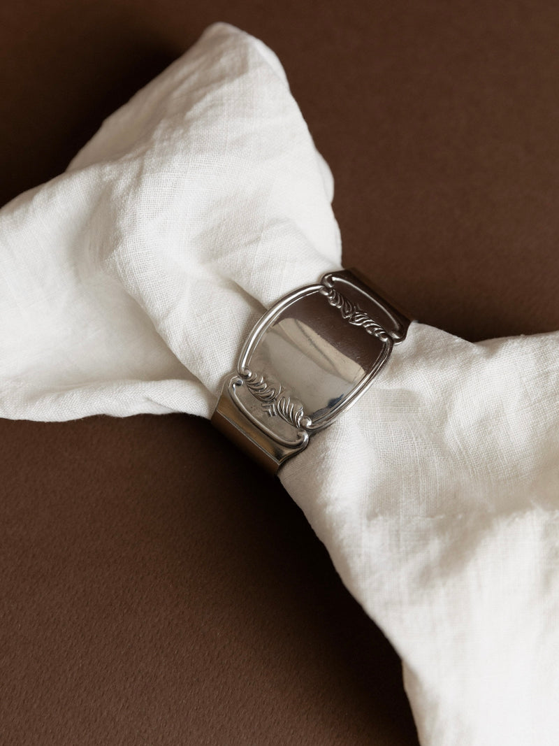 A crisp linen cloth is stylishly wrapped with one of the Set of 6 Stainless Steel Napkin Rings 50s by Bottega Jacobs, featuring a vintage look with embossed leaf designs. This combination, against a warm brown background, adds an exquisite touch to an elegant table setting.