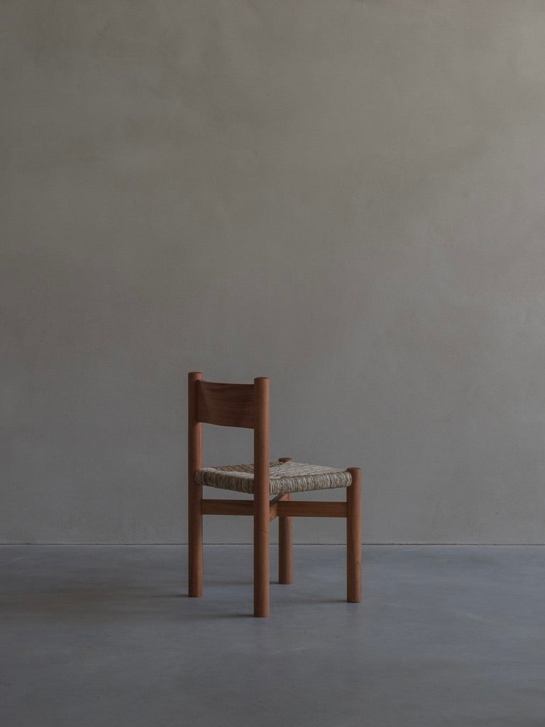 The 'Les Allues' Straw Chair by Bicci de' Medici, crafted from rich mahogany with a woven seat, showcases masterful craftsmanship. It stands on a smooth floor against a muted gray backdrop, featuring simple, clean lines and embodying minimalist design.