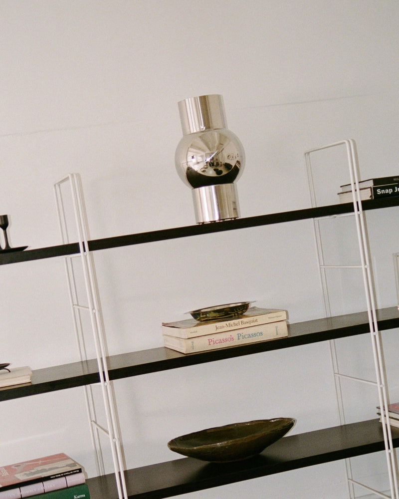 A modern black and white bookshelf with several items. The top shelf holds a reflective stainless steel decorative piece. The middle shelves have books, including "Picasso" and "Picassos," along with the MUHLY-brand Mica Vase in a greenish tone. The lower shelves feature more books, including one with a pink cover.