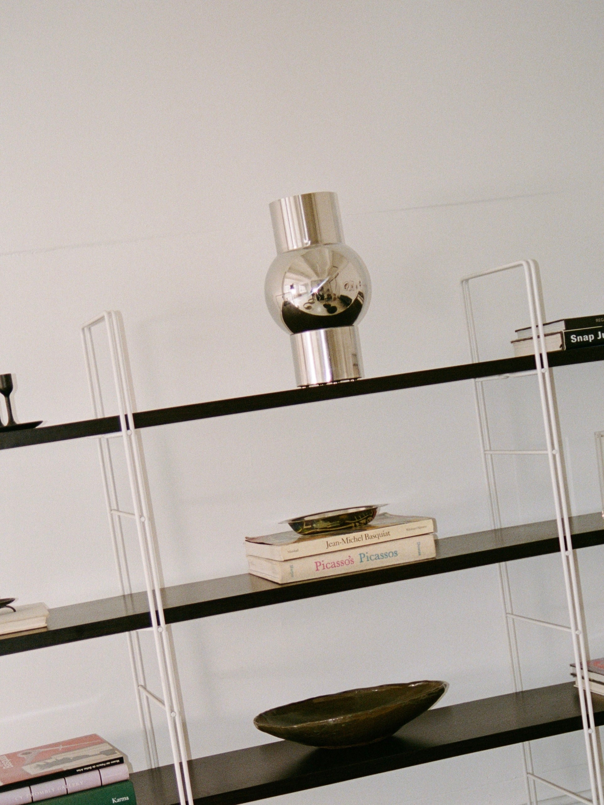 A modern black and white bookshelf with several items. The top shelf holds a reflective stainless steel decorative piece. The middle shelves have books, including "Picasso" and "Picassos," along with the MUHLY-brand Mica Vase in a greenish tone. The lower shelves feature more books, including one with a pink cover.