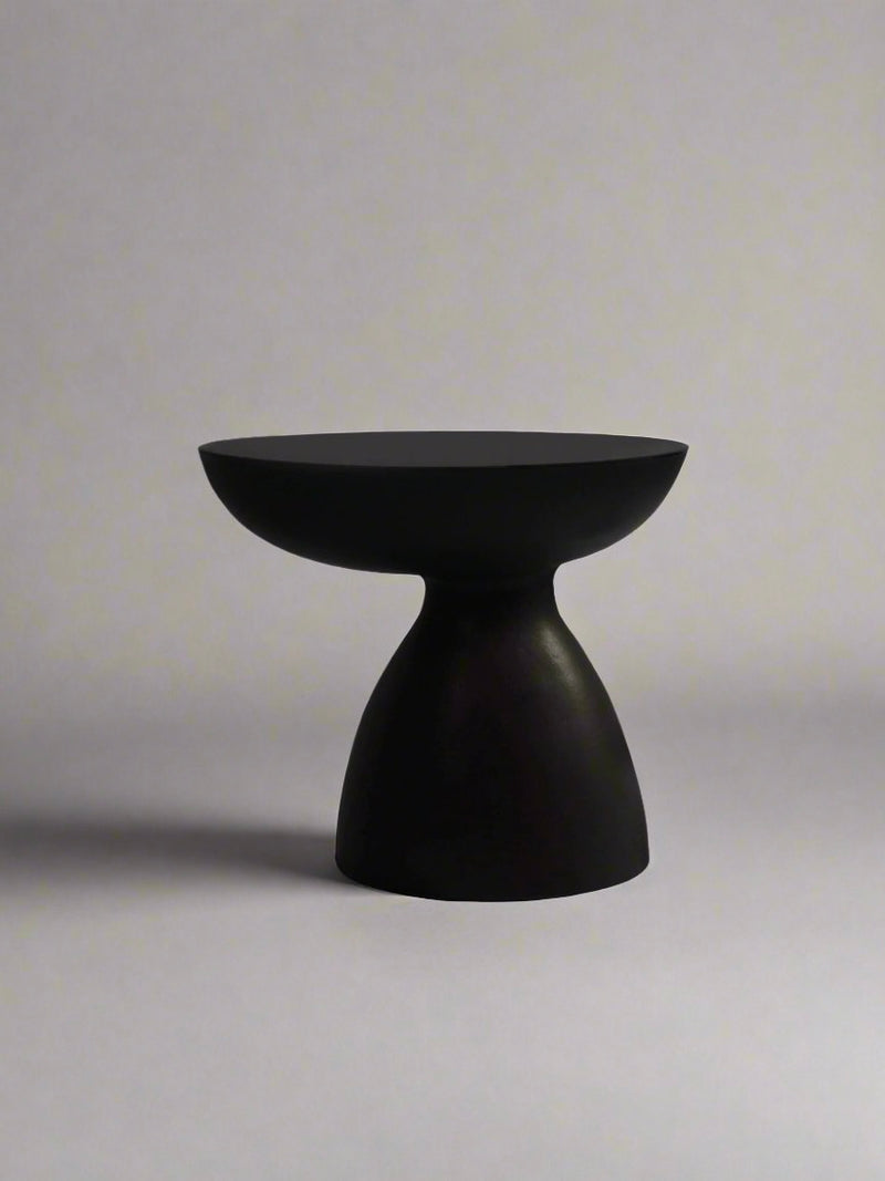 The "Cleo" Ceramic Side Table by Ransom & Dunn is a minimalist black stool with a matte black finish, showcasing a rounded top and a conical base, set against a plain gray background.