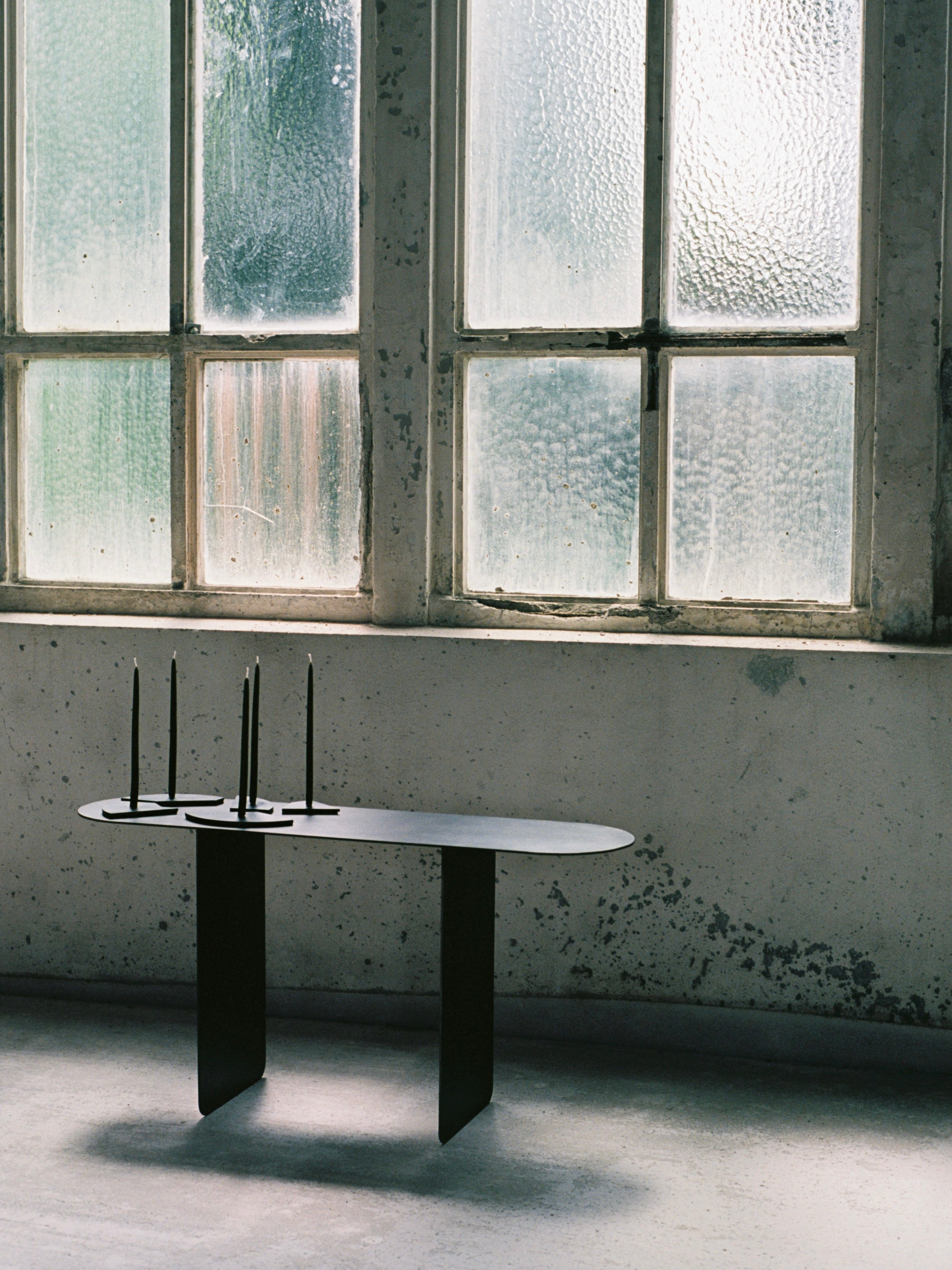 COSEINCORSO's limited edition piece, The Altar, is a modern black table adorned with four unlit black candles. Positioned in front of a textured glass window, it evokes the look of small altars and enhances the minimalist room's industrial aesthetic with its concrete floors and textured wall.