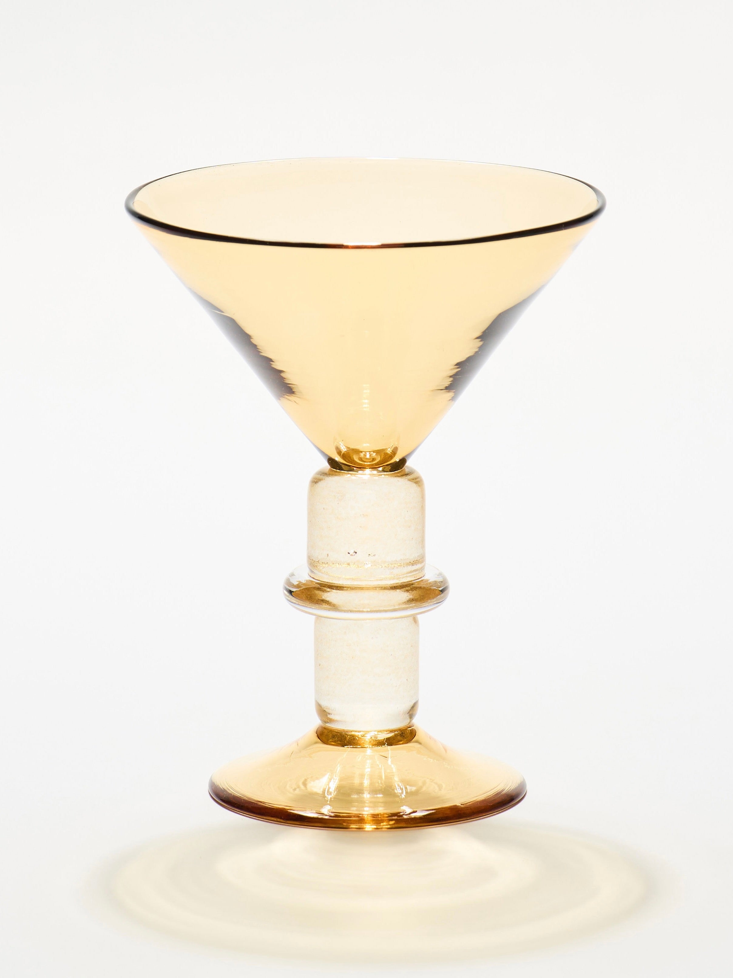 Gather's New York Martini Glass in Whisky boasts a wide, shallow bowl and a sturdy stem with dual ring-like accents. Hand-blown to perfection, this amber masterpiece casts subtle shadows on its simple background.