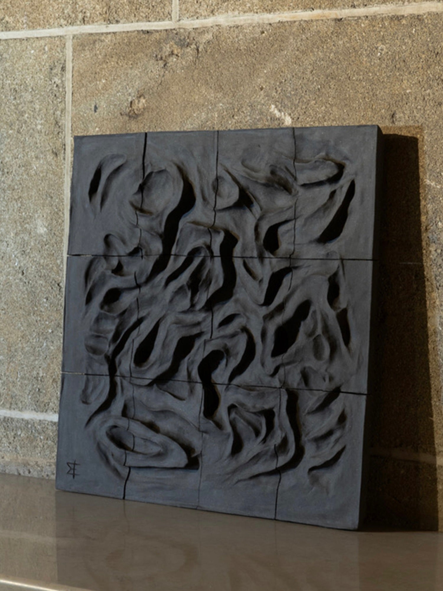 The "Coulée de Lave - Wall Sculpture" by Franck Scala, crafted in textured dark gray ceramic, showcases abstract organic wave-like patterns across multiple square panels forming a larger square. It is elegantly displayed on a shelf against a stone wall.