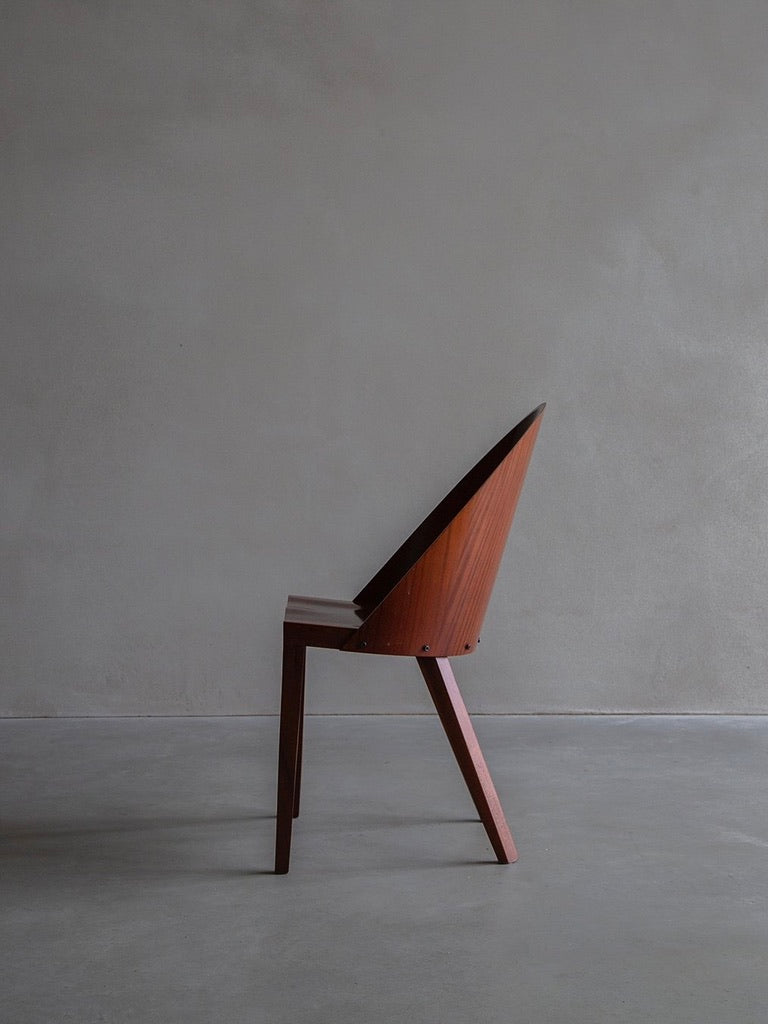 A minimalist wooden chair, the Philippe Starck - Royalton 44 by Bicci de' Medici, with a high triangular backrest and sleek angled legs, sits in a bare room against a textured gray wall. This piece emphasizes clean lines and contemporary aesthetics, reminiscent of Aleph for Driade.