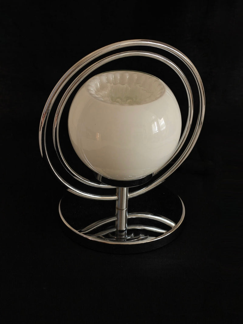 The Space Age Table Lamp by Boga Avante Shop features a spherical glass shade, encircled by concentric spirals of polished chrome metal, all standing on a circular chrome base against a black background.
