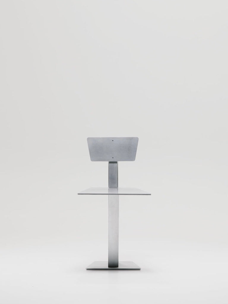 A sleek, minimalistic metal stand with a rectangular base and flat top is set against a plain white background. The design mirrors the modern style of Marquel Williams' handcrafted Beam Chairs, featuring clean lines and a silver finish.