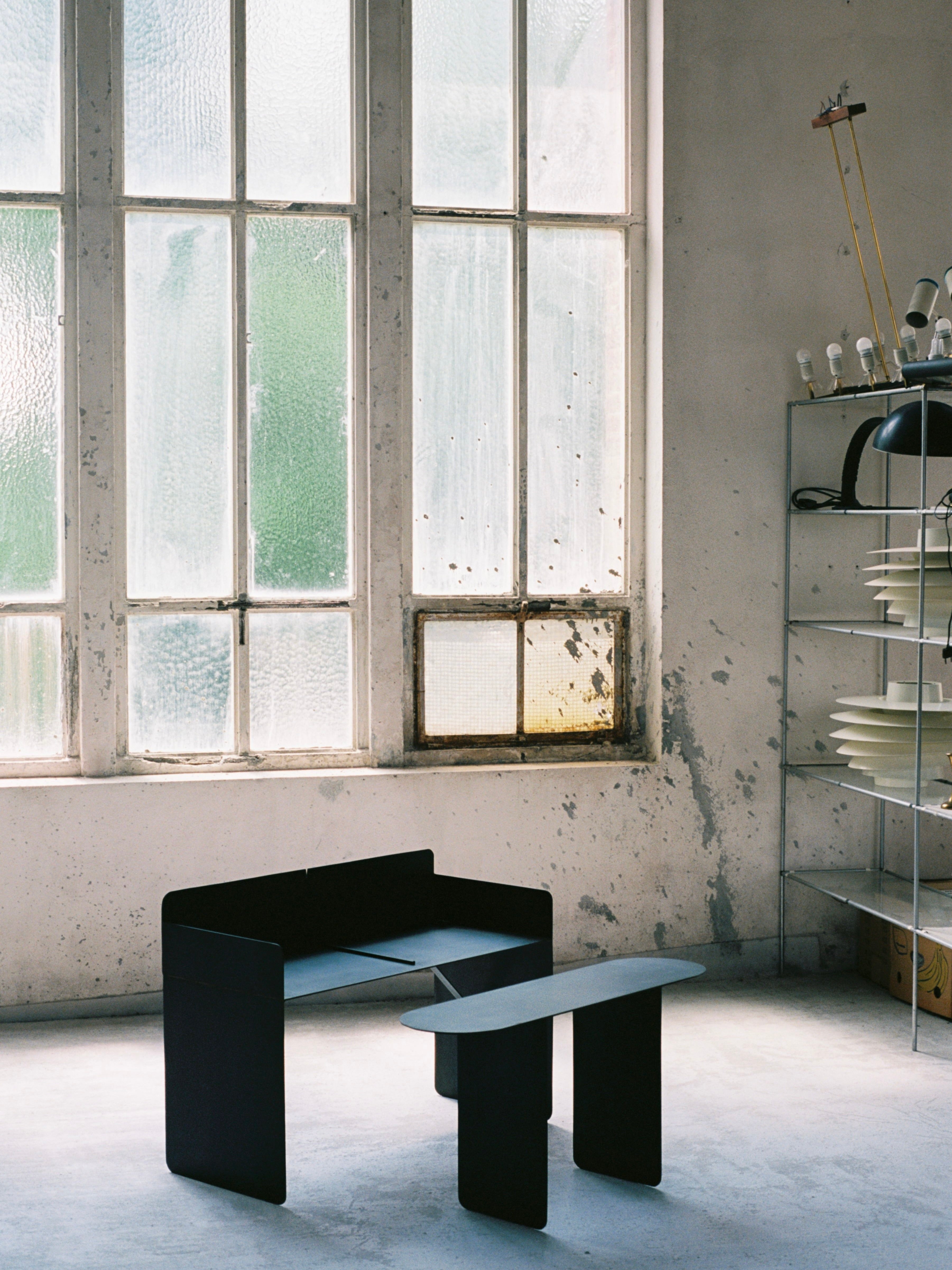 A modern, minimalistic interior features "The Altar," a black table and bench set from COSEINCORSO, placed against large textured windows. The room exudes an industrial vibe with a metal shelving unit displaying various objects alongside limited edition pieces, as soft, natural light filters through the windows.