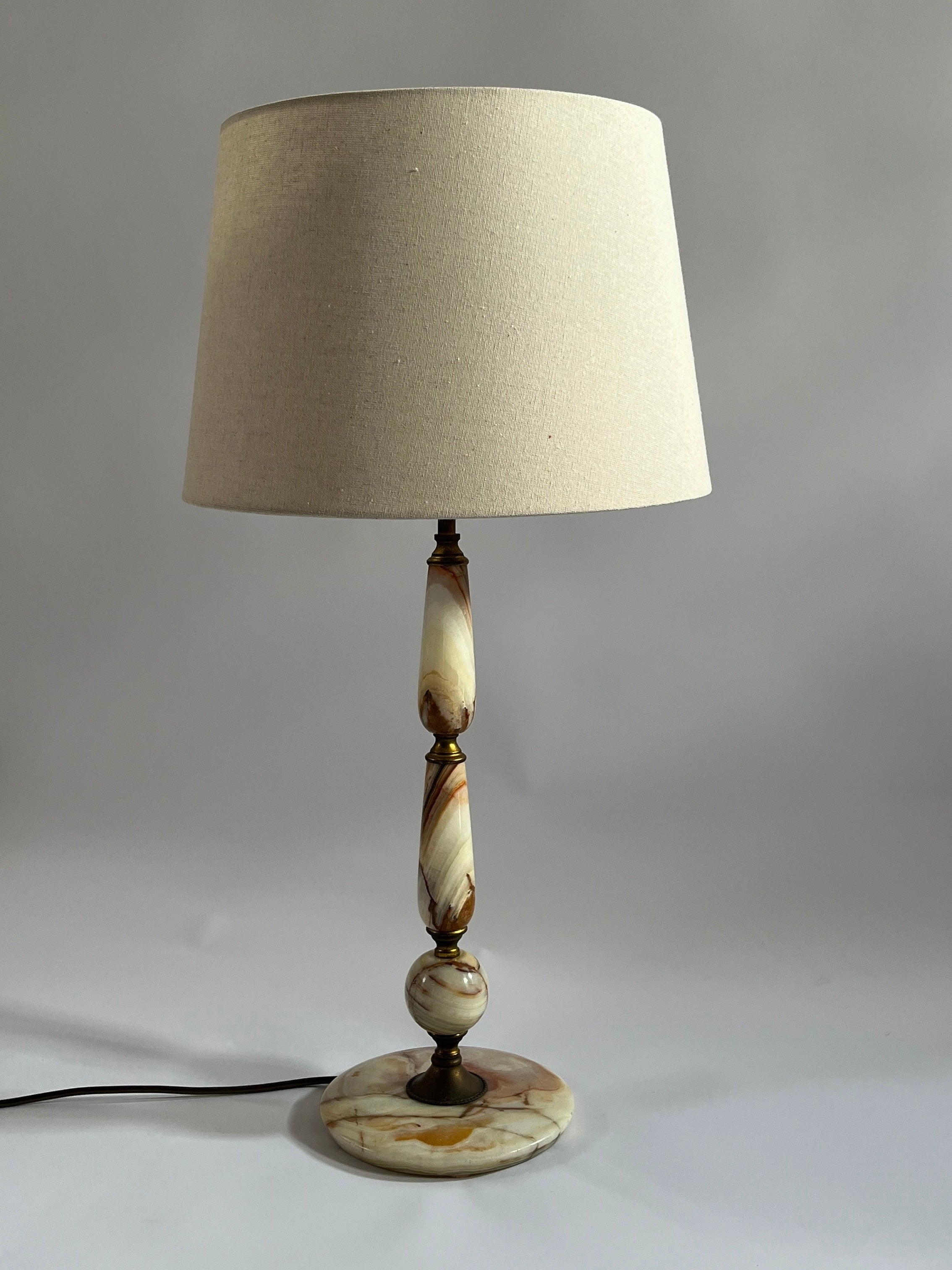 The Maud Vaughan Vintage Onyx Table Lamp features a beige lampshade and a base adorned with marbled swirls of beige, cream, and brown. The elegant base consists of a circular bottom, two elongated and ornate sections accented with brass details, and a thin central rod, creating a timeless classic look.