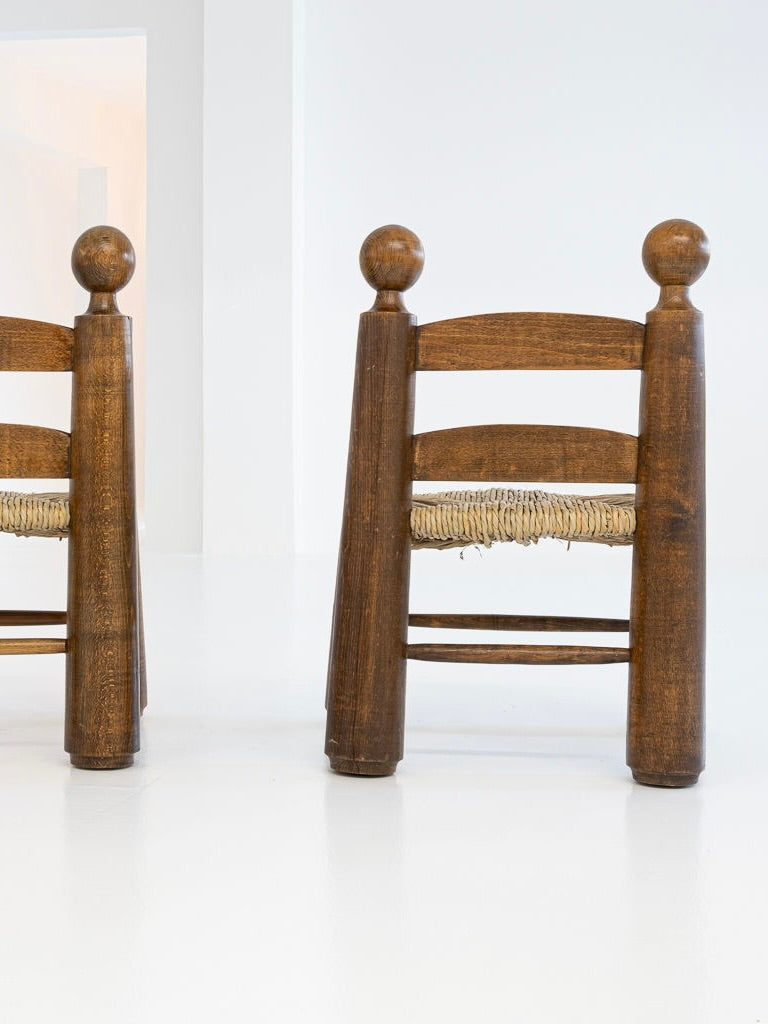 This set includes two solid oak chairs with a rustic ladder-back design, round finials, and woven seats, reminiscent of Charles Dudouyt's style. The simple white background enhances their minimalist appeal. This item is by antibeige and also includes one table from the 1940s.