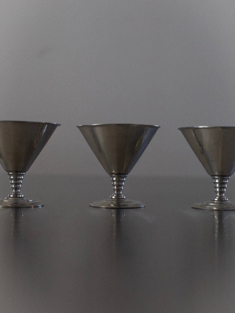 A photograph features three elegant ice cream cups from the "Six Ice Cream Cups" collection by Out For Lunch, arranged in a line on a reflective surface. Each cup is adorned with a wide, flared rim and rests on a short, sturdy stem. The plain gray background enhances the minimalist and sophisticated appeal of these Norwegian-inspired goblets.