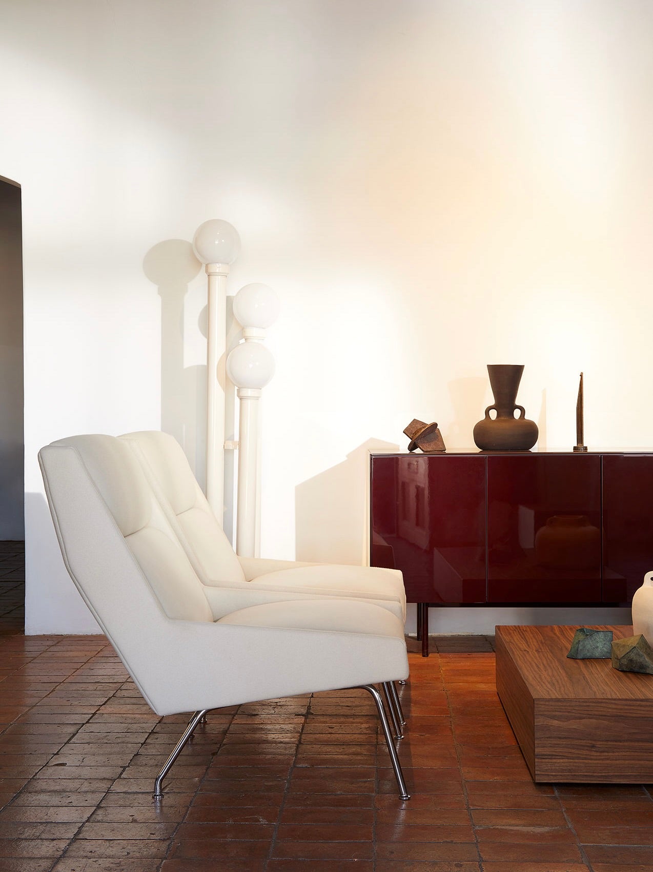 A modern living room features two "Kent" Armchairs by Lemon, upholstered in cotton velvet, alongside a dark red sideboard displaying decorative vases. The room includes a wooden coffee table and a distinctive floor lamp with multiple globes. With terracotta flooring, the space exudes a warm, minimalist aesthetic inspired by Italian 1950s design.