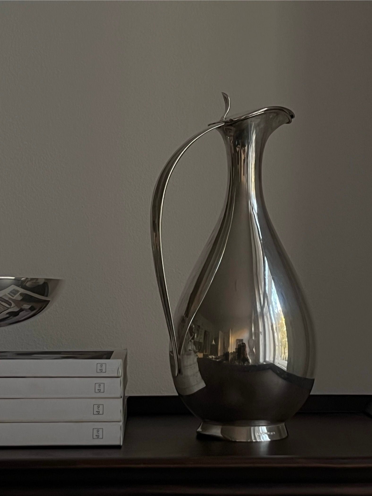 The Mid-Century Silver Water Pitcher by Médecine, with its sleek metallic design inspired by rare 1950s Finnish water jugs, rests on a dark surface next to three stacked white books and a silver bowl, all framed by a plain, light-colored wall.